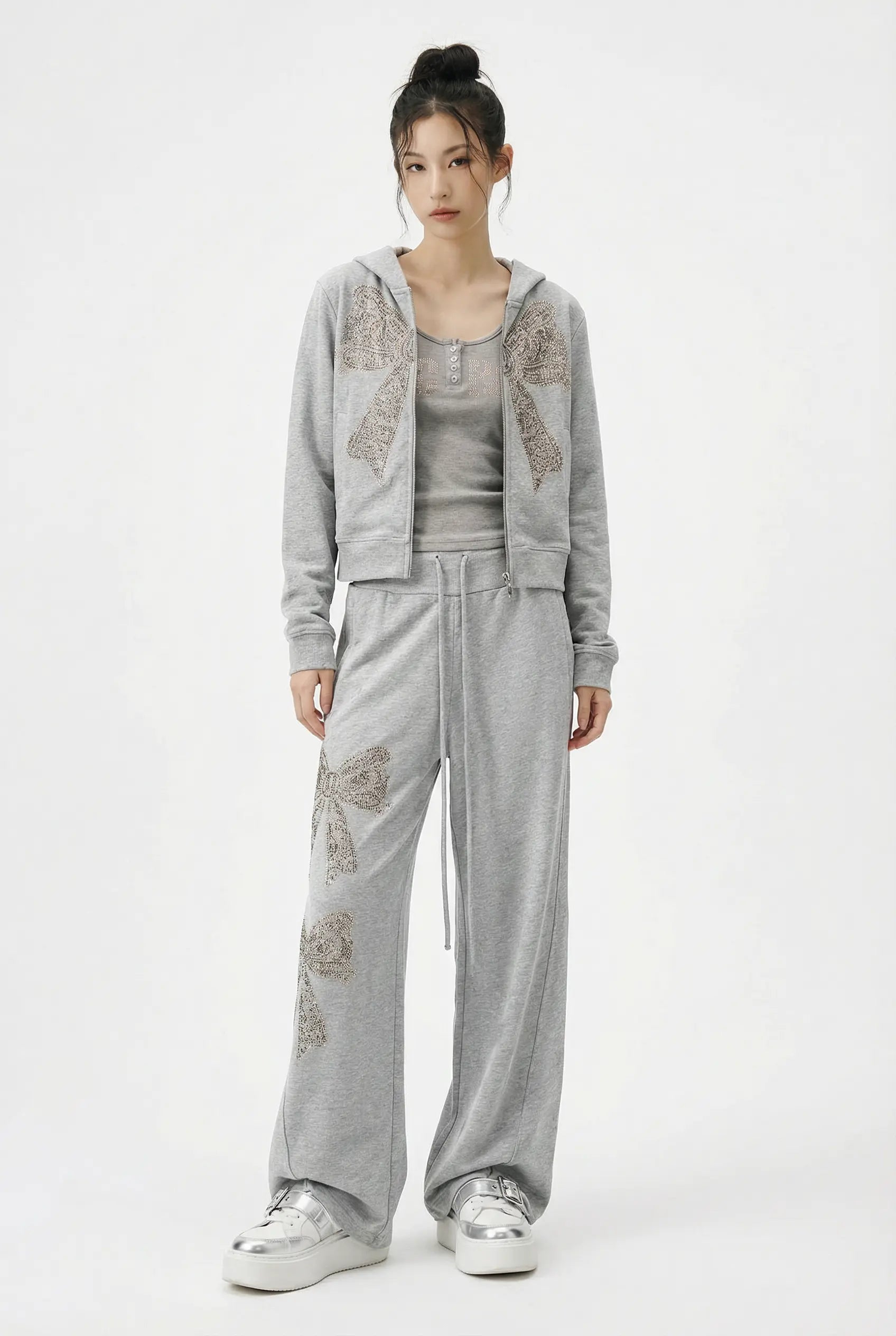 A model wearing a light gray two-piece loungewear set featuring a cropped zip-up hoodie with beige floral embroidery on the sleeves and matching wide-leg sweatpants, paired with a gray tank top and white sneakers. TCH Rhinestone Bow 3-Piece Lounge Set