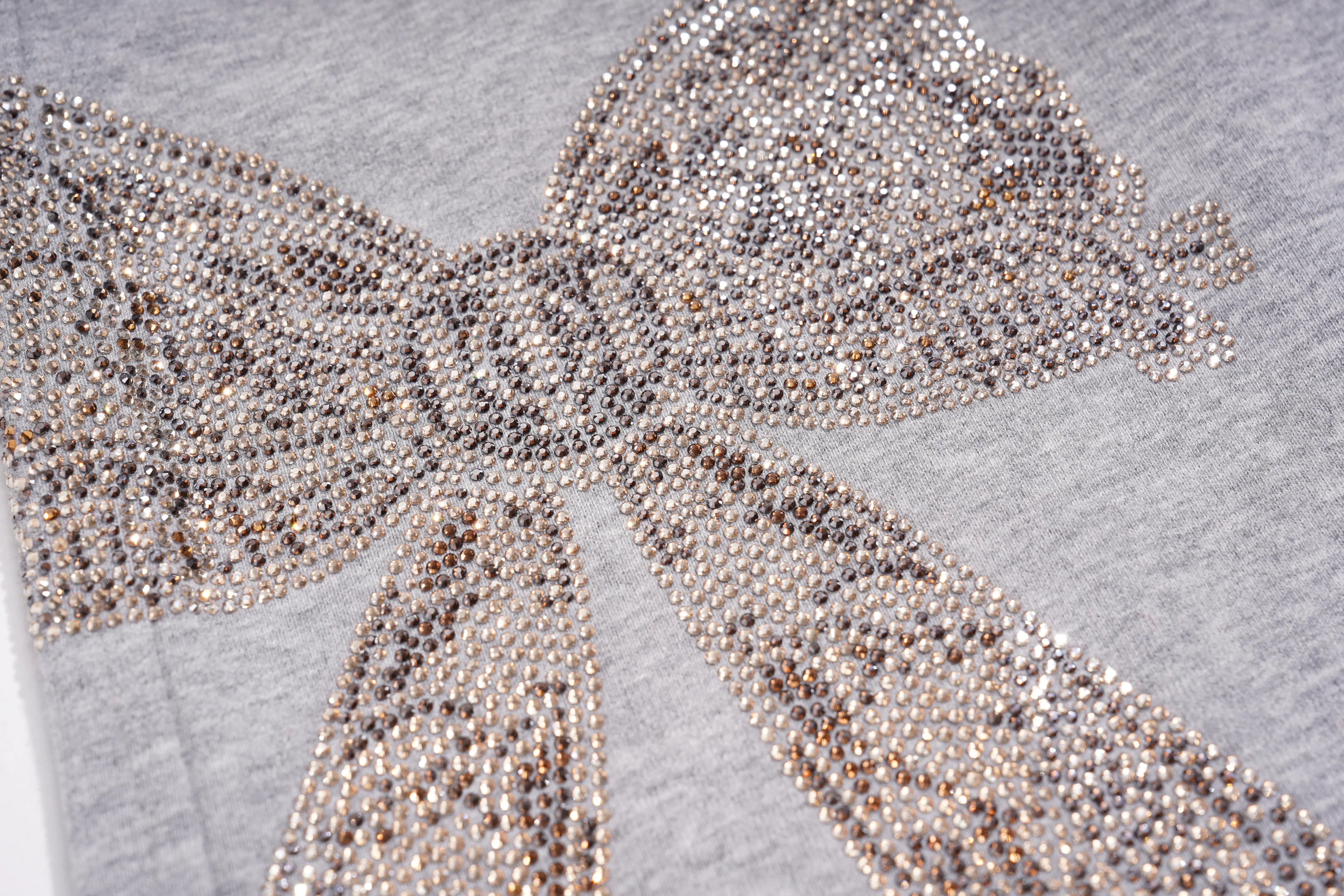 A close-up of a gray fabric item decorated with a large bow design made of dense rhinestones in silver, champagne, and rose gold tones. TCH Rhinestone Bow 3-Piece Lounge Set