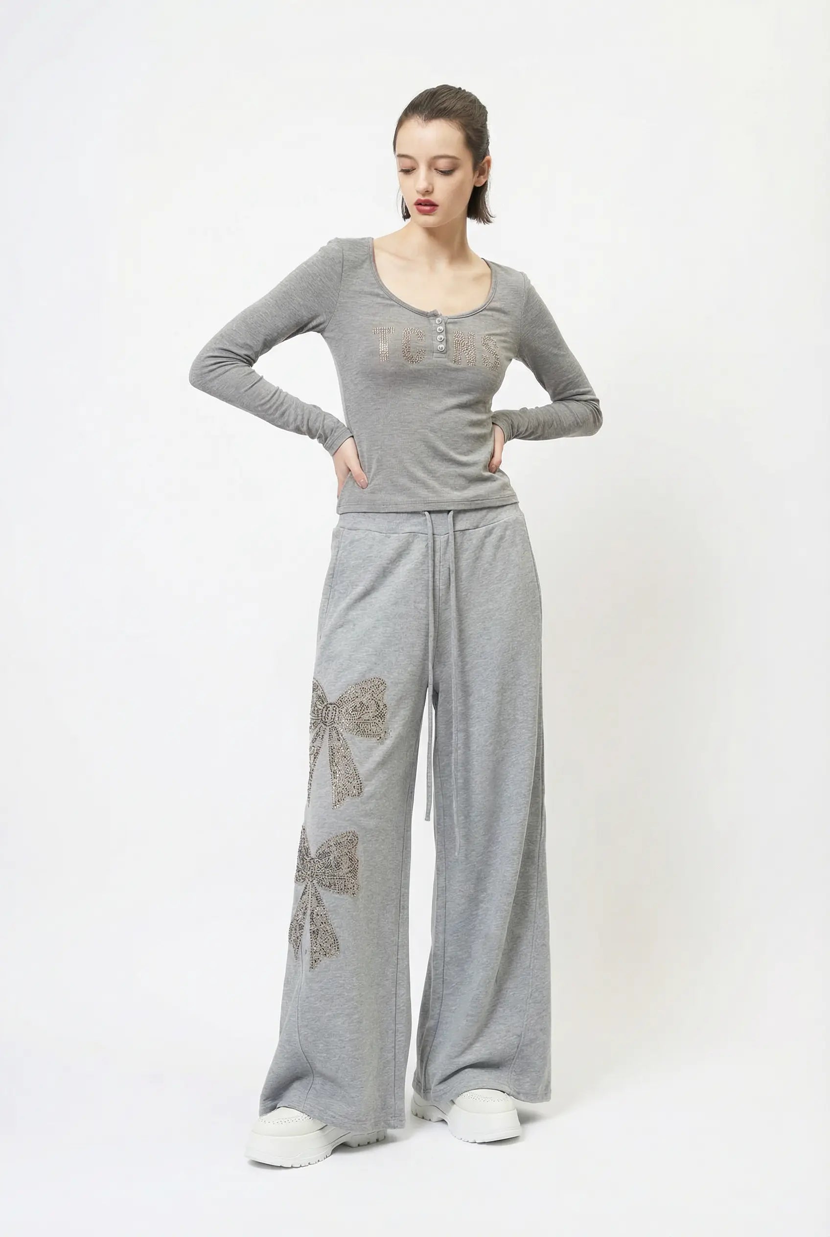 A woman modeling a gray long-sleeve henley top and matching wide-leg sweatpants with bow embellishments, standing against a plain white studio background. TCH Rhinestone Bow 3-Piece Lounge Set