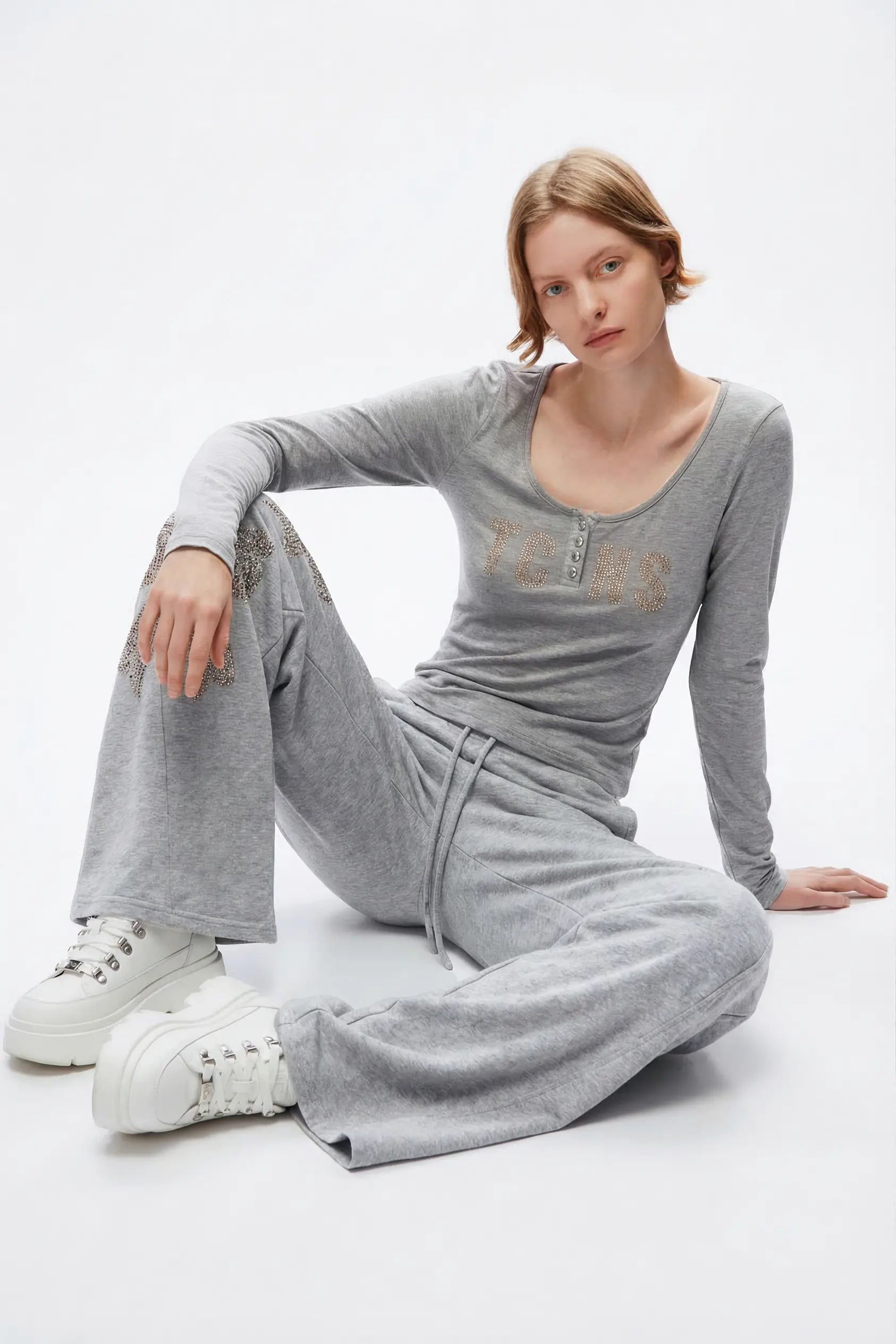 A woman wearing a light gray two-piece loungewear set consisting of a long-sleeve henley top with gold lettering and matching wide-leg sweatpants, sitting on a white studio floor. TCH Rhinestone Bow 3-Piece Lounge Set