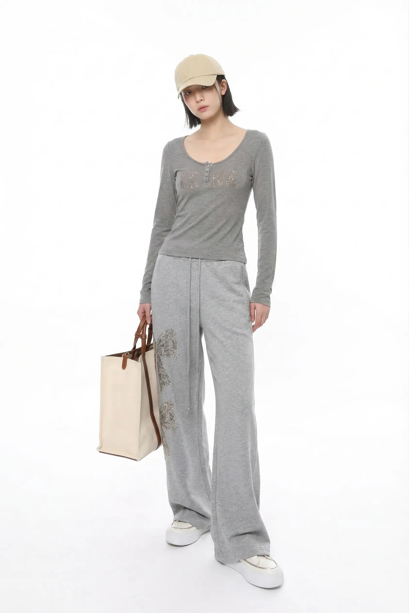 A model wearing a gray long-sleeve top with lace detailing, matching gray wide-leg sweatpants with floral embroidery, a beige baseball cap, and white sneakers, holding a beige tote bag against a white background. TCH Rhinestone Bow 3-Piece Lounge Set