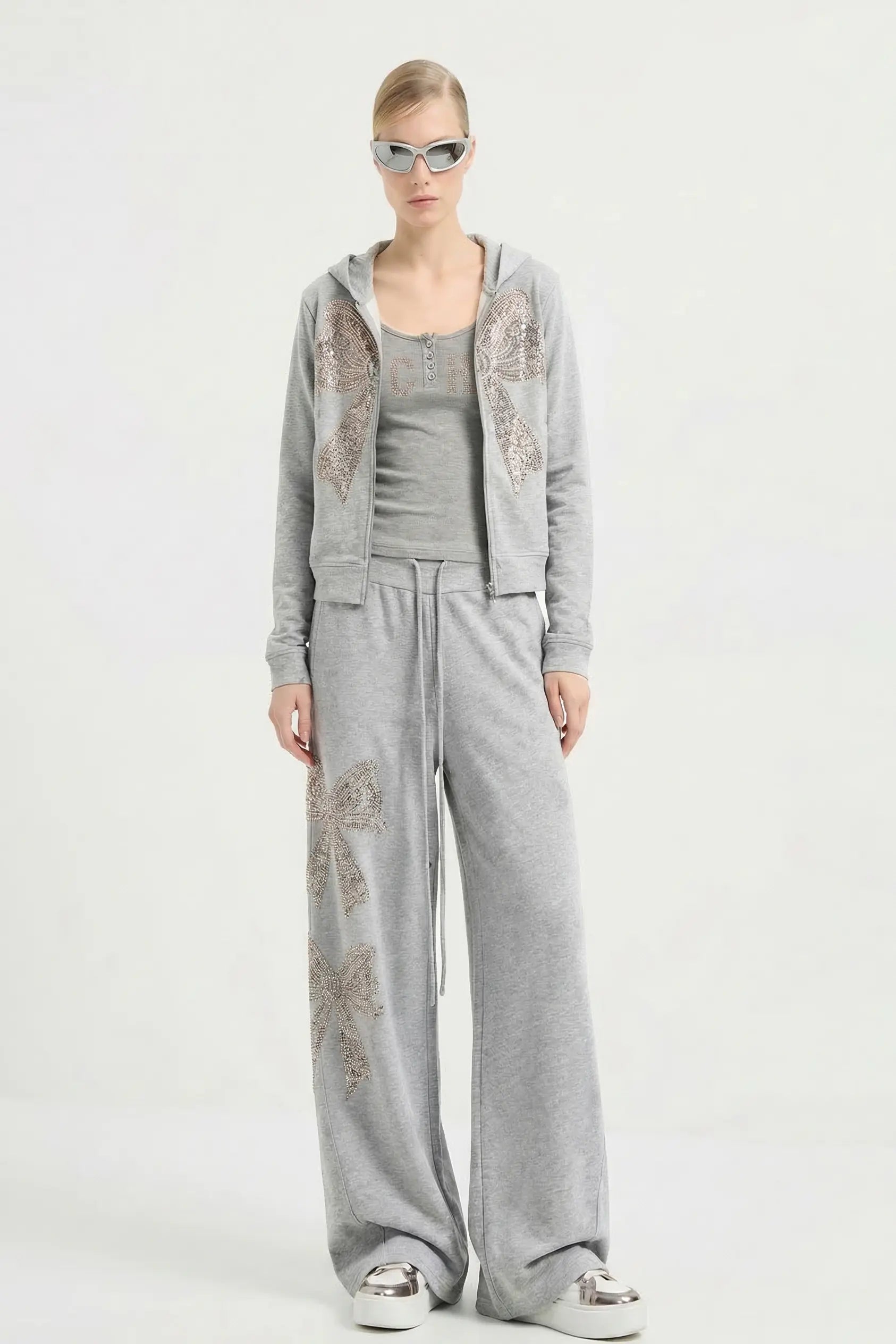 A model wearing a light gray two-piece loungewear set featuring a cropped zip-up hoodie with butterfly embellishments and matching wide-leg sweatpants, styled with sunglasses and sneakers against a plain white studio background. TCH Rhinestone Bow 3-Piece Lounge Set
