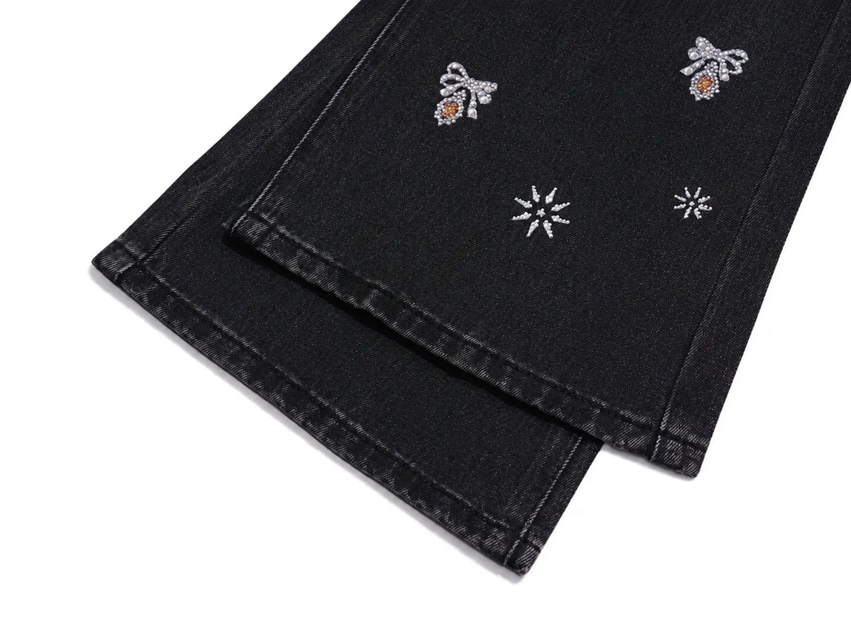 Close-up of black denim jeans folded over, showing embroidered details including two silver bows with orange centers and two silver snowflake motifs on a white background. TCH Rhinestone Bow & Butterfly Flare Jeans