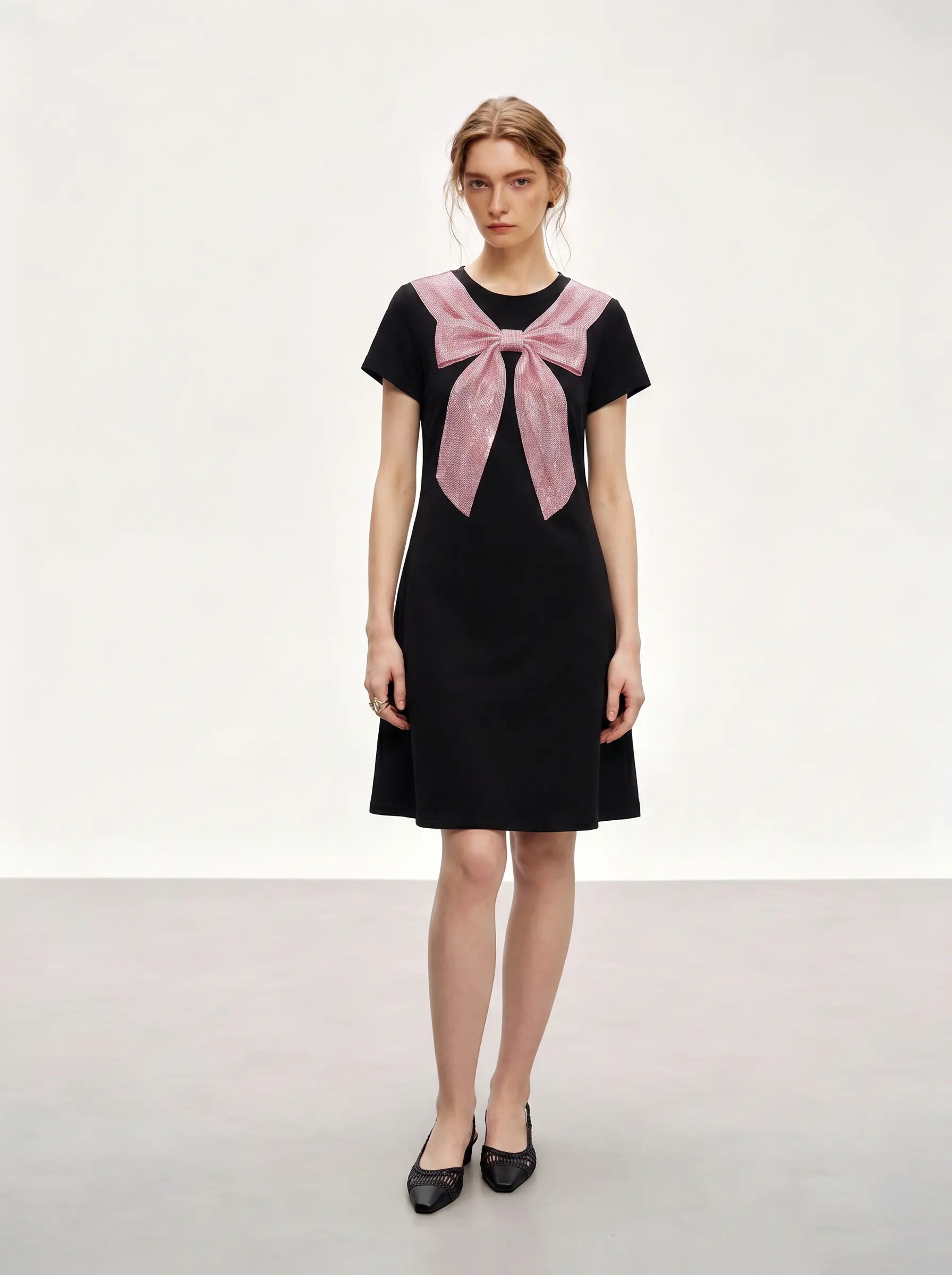 A woman modeling a black short-sleeve knee-length dress with a large pink bow detail at the neckline, paired with black pointed flats, against a plain light gray studio background. TCH Rhinestone Bow Collar Black Mini Dress
