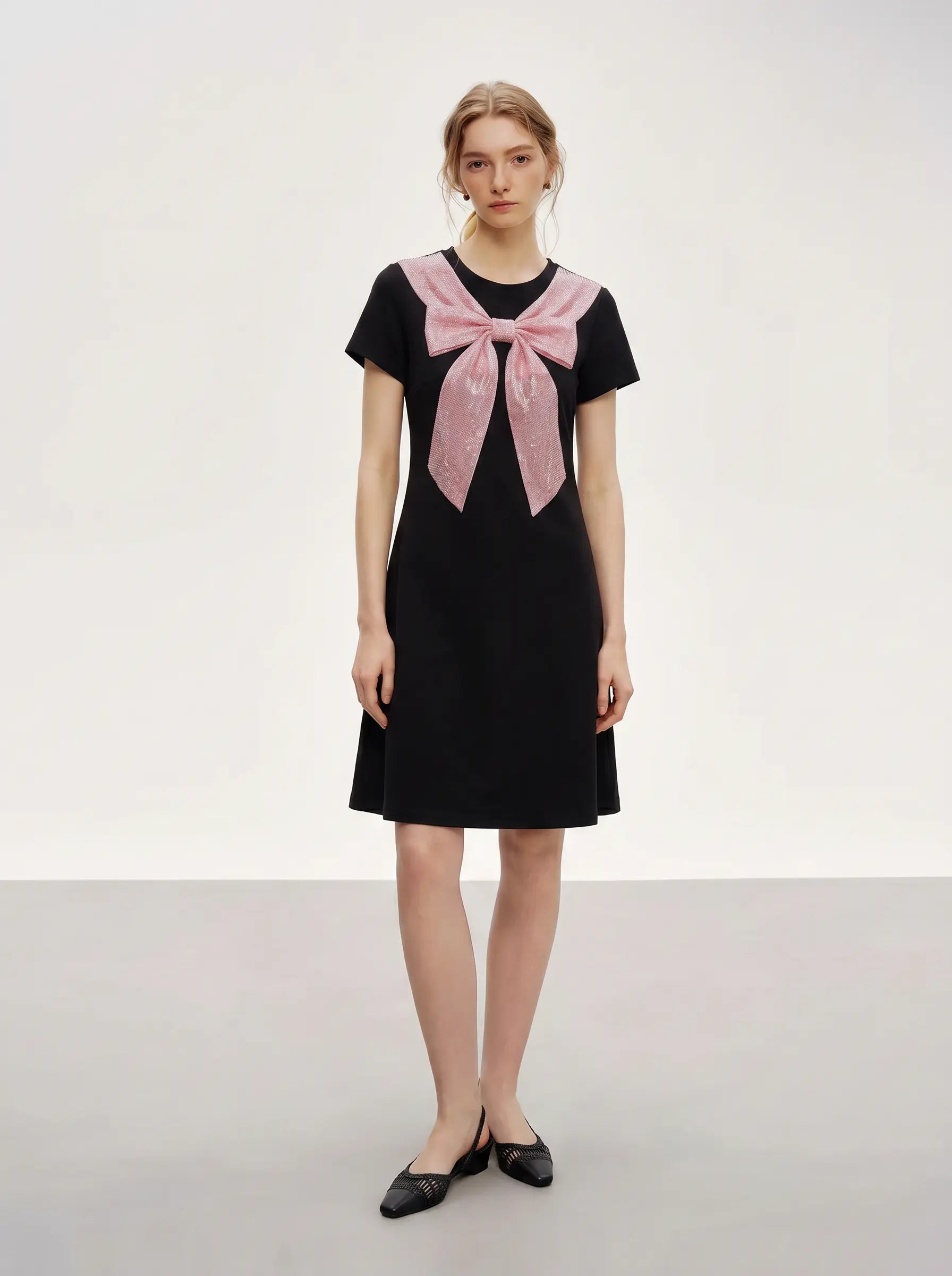 A woman modeling a black short-sleeve dress with a large pink bow detail at the neckline, paired with black pointed flats, against a plain light gray studio background. TCH Rhinestone Bow Collar Black Mini Dress