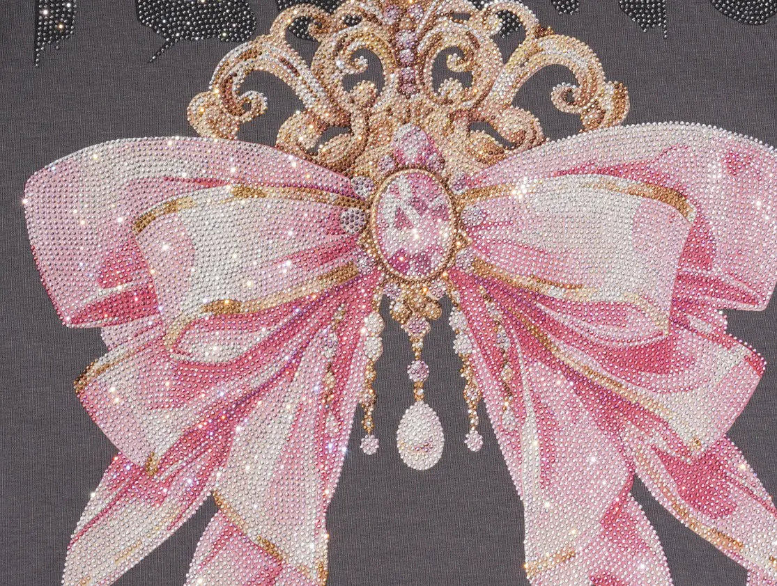 A close-up of a pink and white beaded bow embellishment with gold-toned scrollwork and dangling rhinestone accents on a dark gray background. TCH Rhinestone Bow Crown Graphic Mini Dress