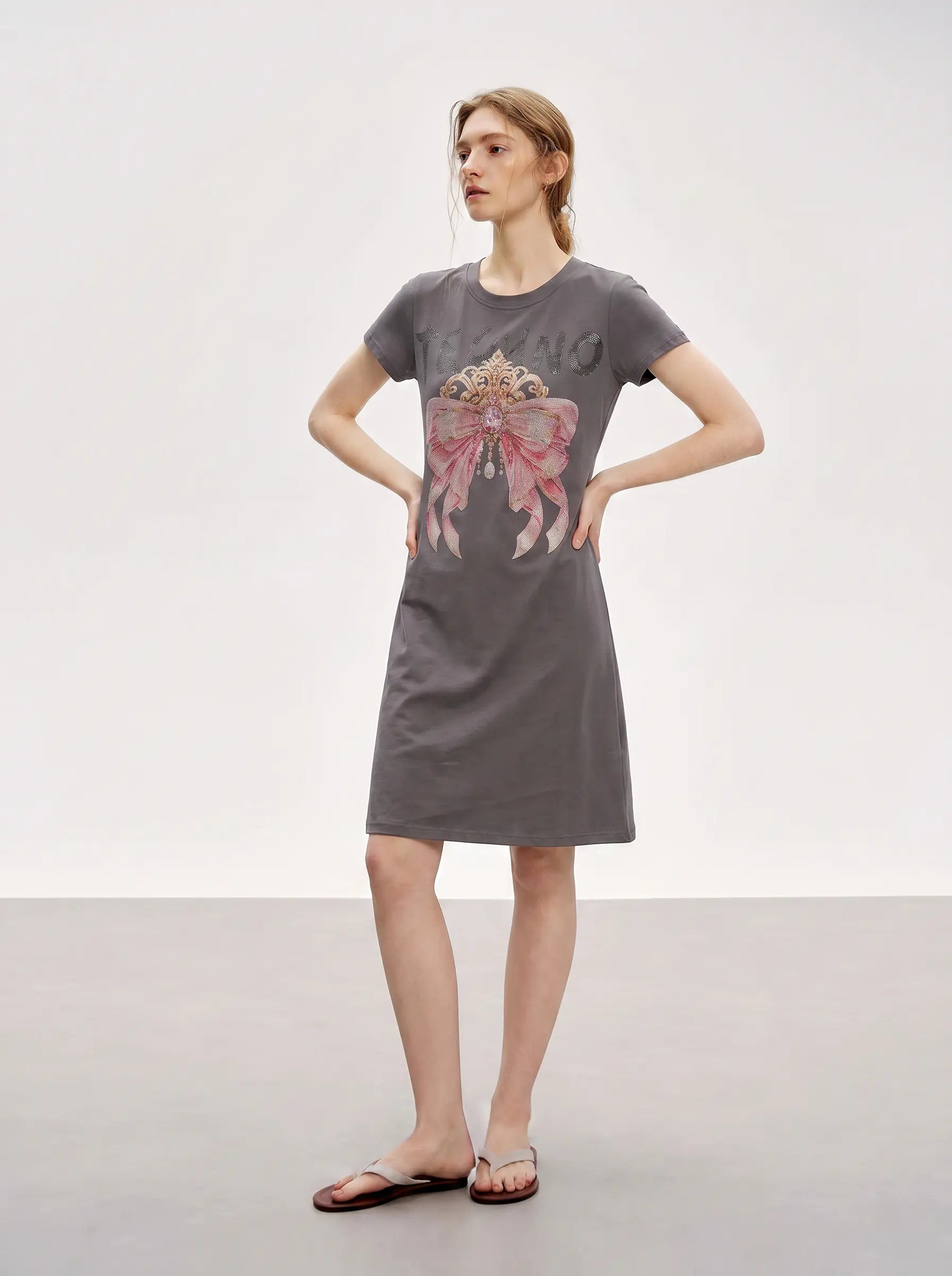 A woman modeling a dark gray short-sleeve t-shirt dress with a large pink bow graphic on the front, paired with brown flip-flop sandals, set against a plain light gray studio background. TCH Rhinestone Bow Crown Graphic Mini Dress