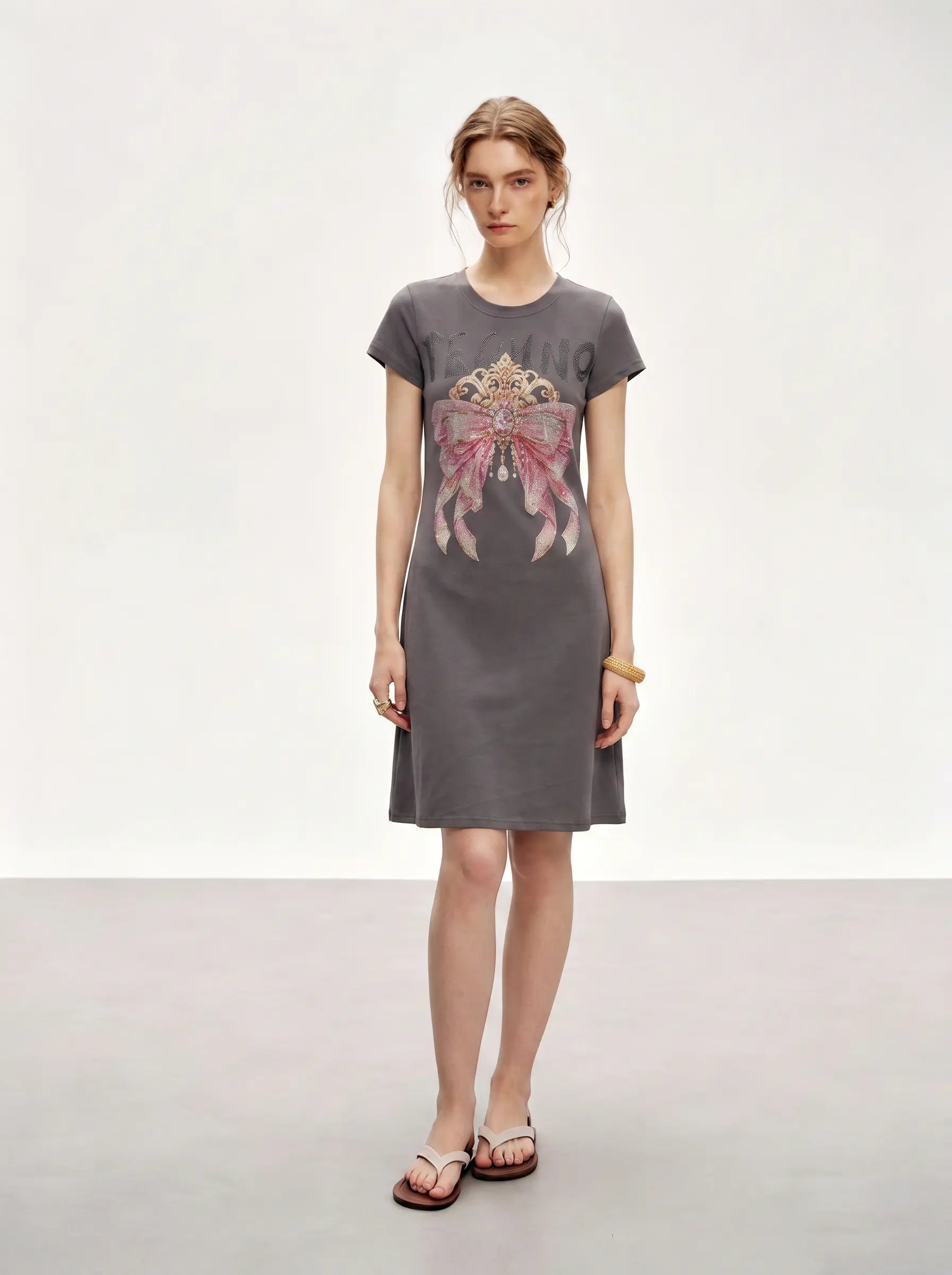 A woman modeling a short-sleeve gray knit dress with a large pink and gold graphic print on the front, paired with brown sandals, against a plain light gray studio background. TCH Rhinestone Bow Crown Graphic Mini Dress