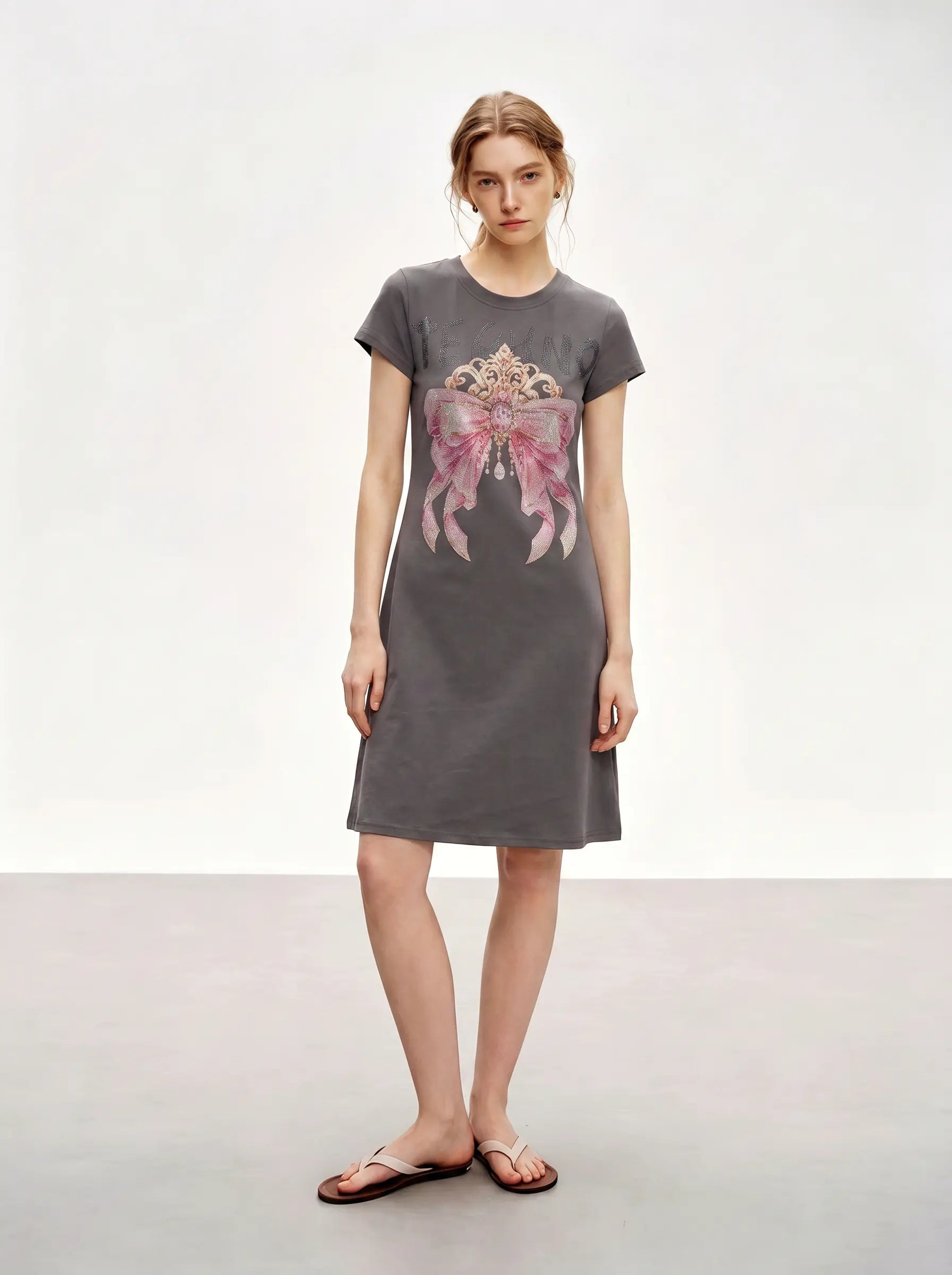 A woman modeling a dark gray short-sleeve t-shirt dress with a large pink bow graphic on the front, paired with brown flip-flop sandals, set against a plain light gray studio background. TCH Rhinestone Bow Crown Graphic Mini Dress