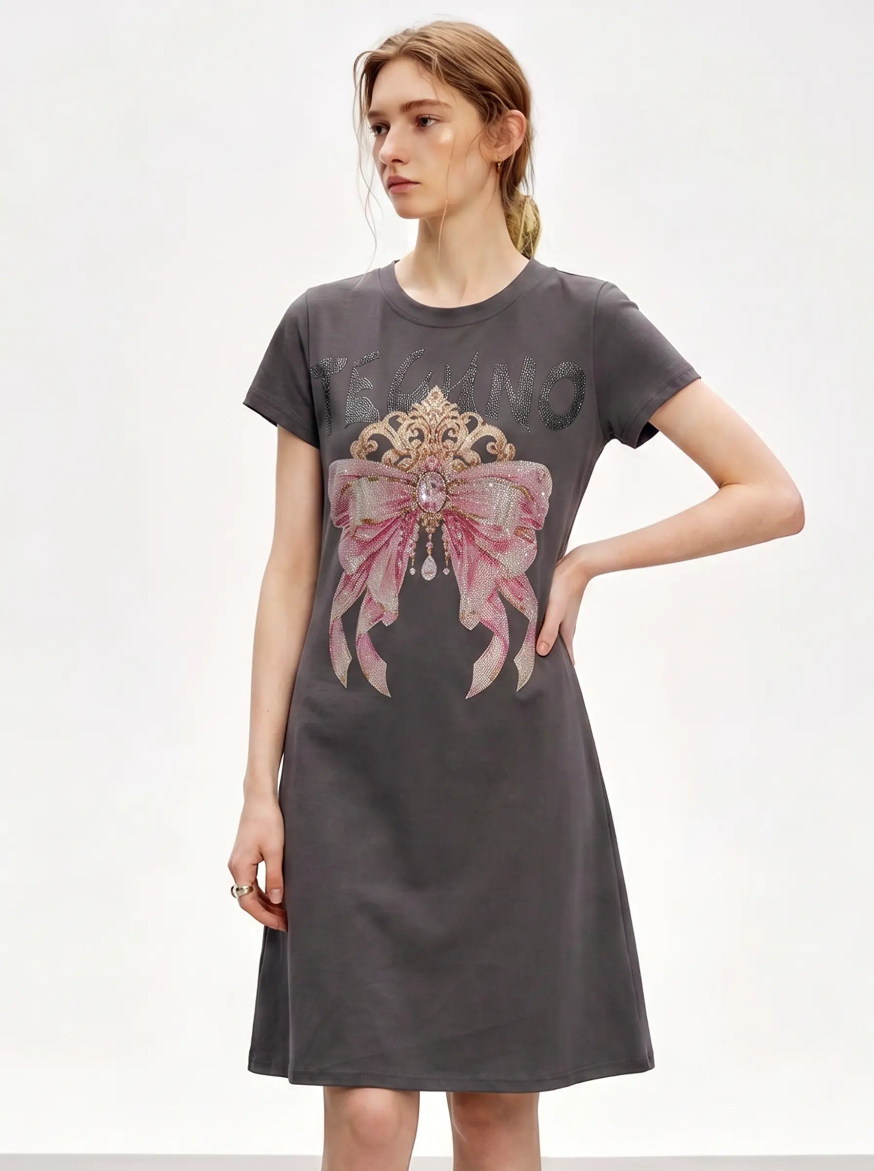 A woman modeling a dark gray short-sleeve t-shirt dress with a large pink and gold bow graphic on the front, set against a plain white studio background. TCH Rhinestone Bow Crown Graphic Mini Dress
