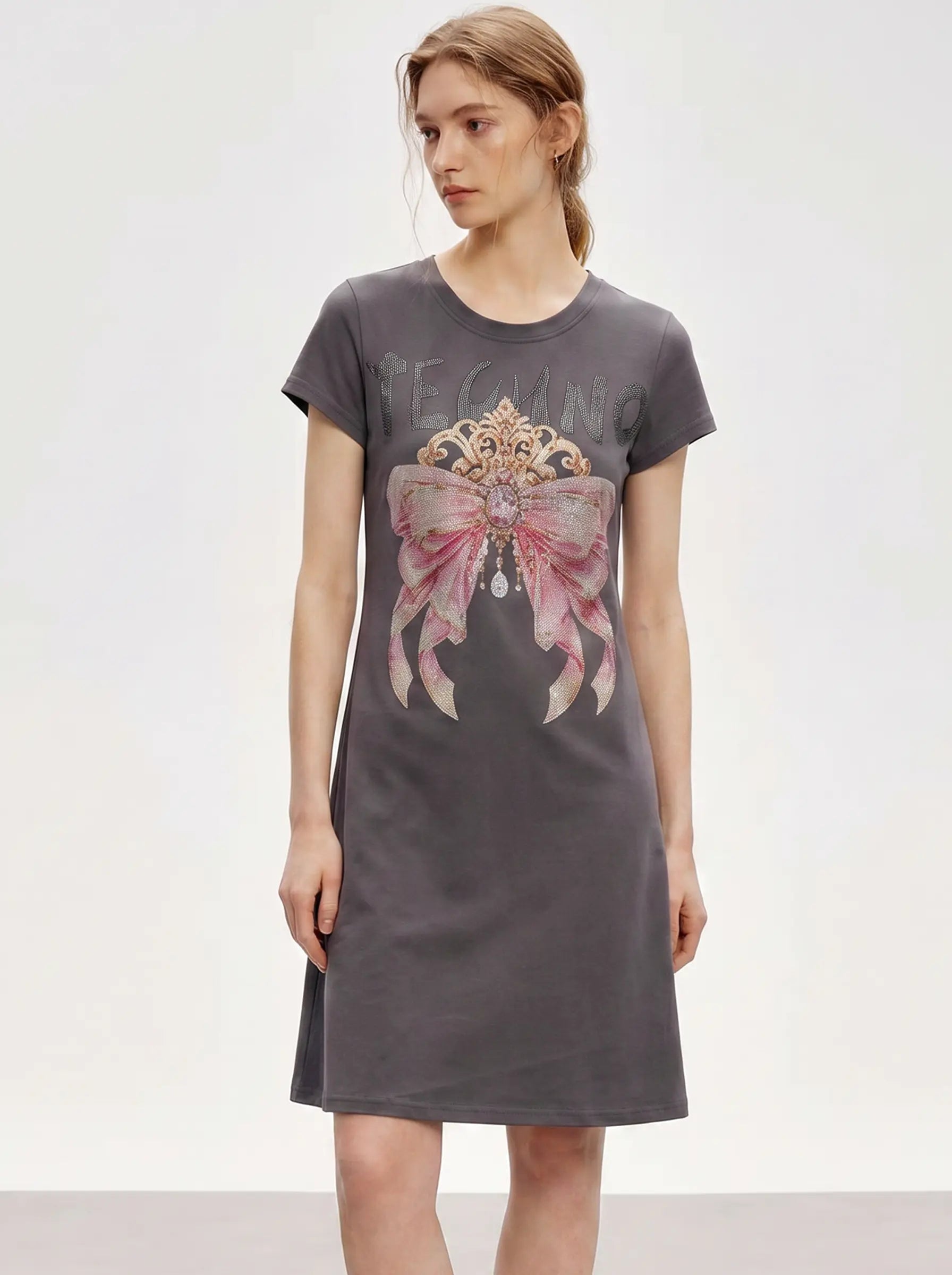 A woman modeling a dark gray short-sleeve t-shirt dress with a large graphic print of a pink bow and a gold crown on the front, set against a plain light gray studio background. TCH Rhinestone Bow Crown Graphic Mini Dress