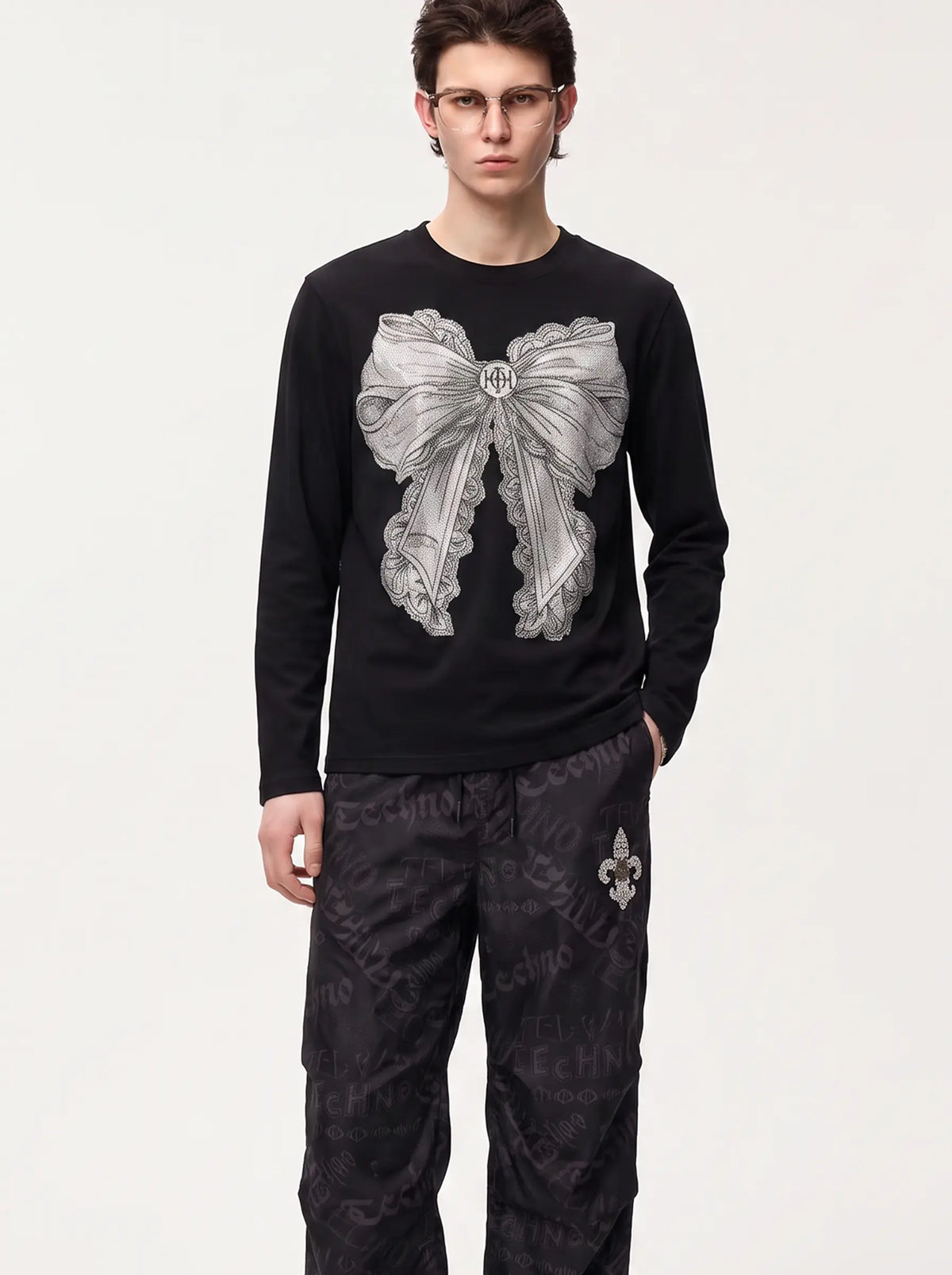 A model wearing a black long-sleeve shirt with a large silver bow graphic on the front, paired with dark patterned pants, against a plain light gray background. TCH Rhinestone Bow Graphic Fitted Long Sleeve Top