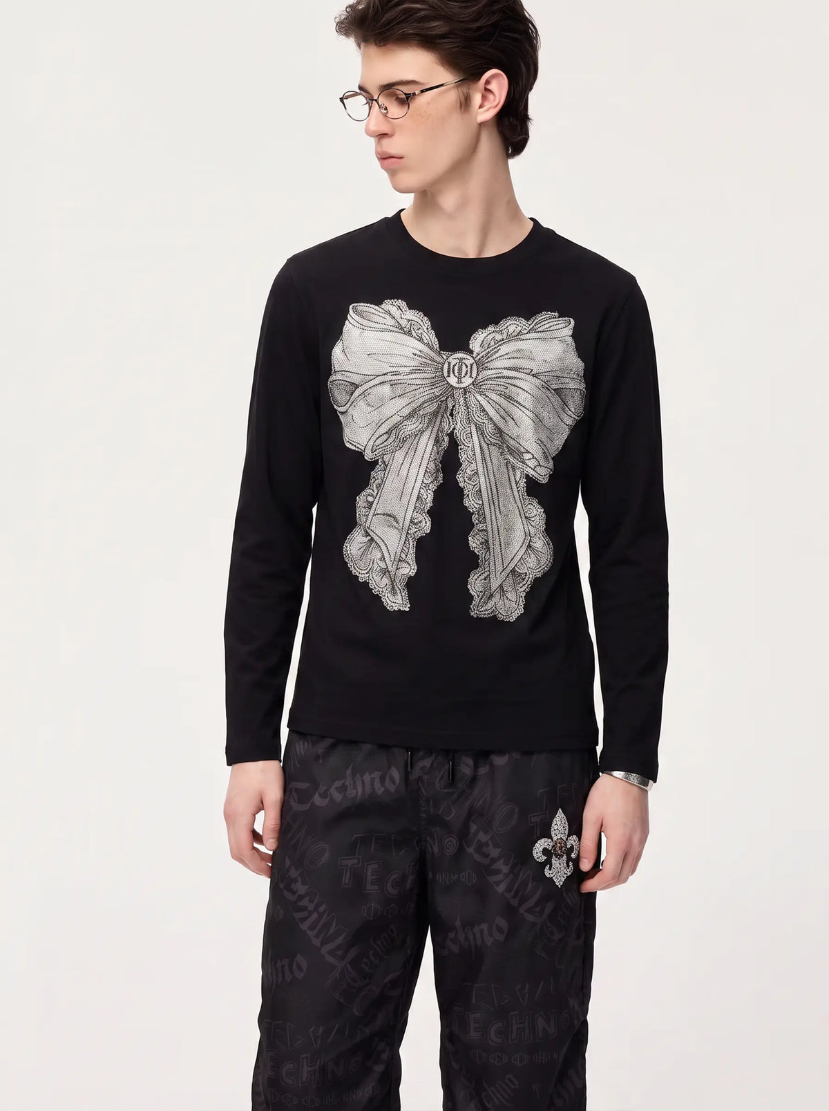 A model wearing a black long-sleeve crewneck shirt with a large gray bow graphic on the front, paired with black patterned pants, against a plain light background. TCH Rhinestone Bow Graphic Fitted Long Sleeve Top
