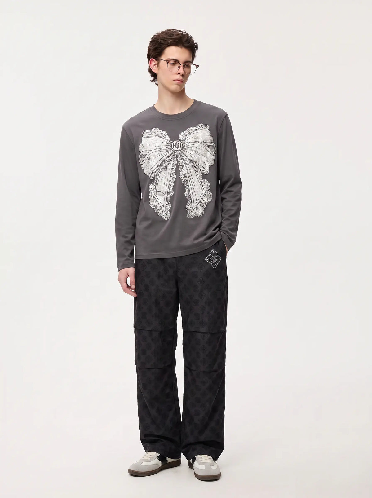 A man wearing a dark gray long-sleeve t-shirt with a large white bow graphic on the front, paired with dark patterned cargo pants and white sneakers, standing against a plain light gray studio background. TCH Rhinestone Bow Graphic Fitted Long Sleeve Top