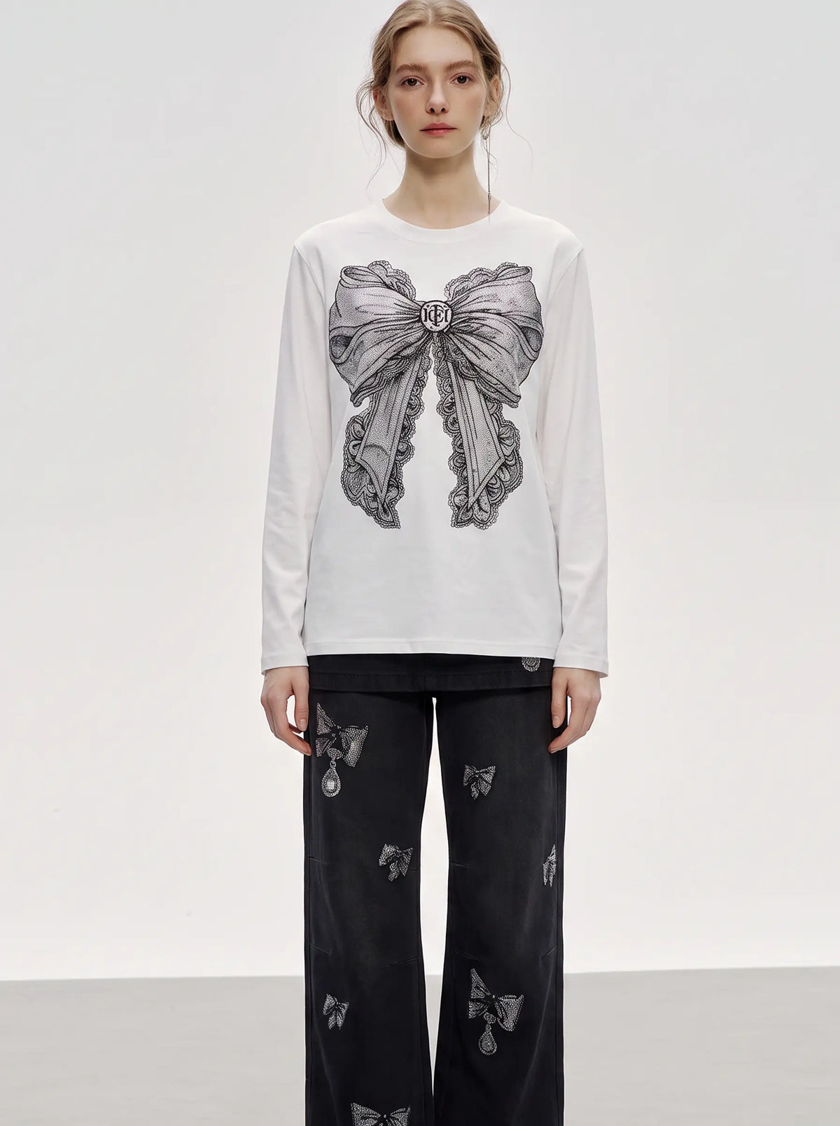 A model stands against a light gray studio background wearing a white long-sleeve top with a large gray bow graphic on the front, paired with black wide-leg pants featuring small bow motifs. TCH Rhinestone Bow Graphic Fitted Long Sleeve Top