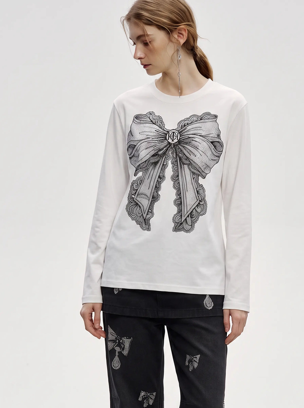 A woman modeling a white long-sleeve top with a large black and gray bow graphic on the front, paired with dark patterned pants, against a plain light gray studio background. TCH Rhinestone Bow Graphic Fitted Long Sleeve Top