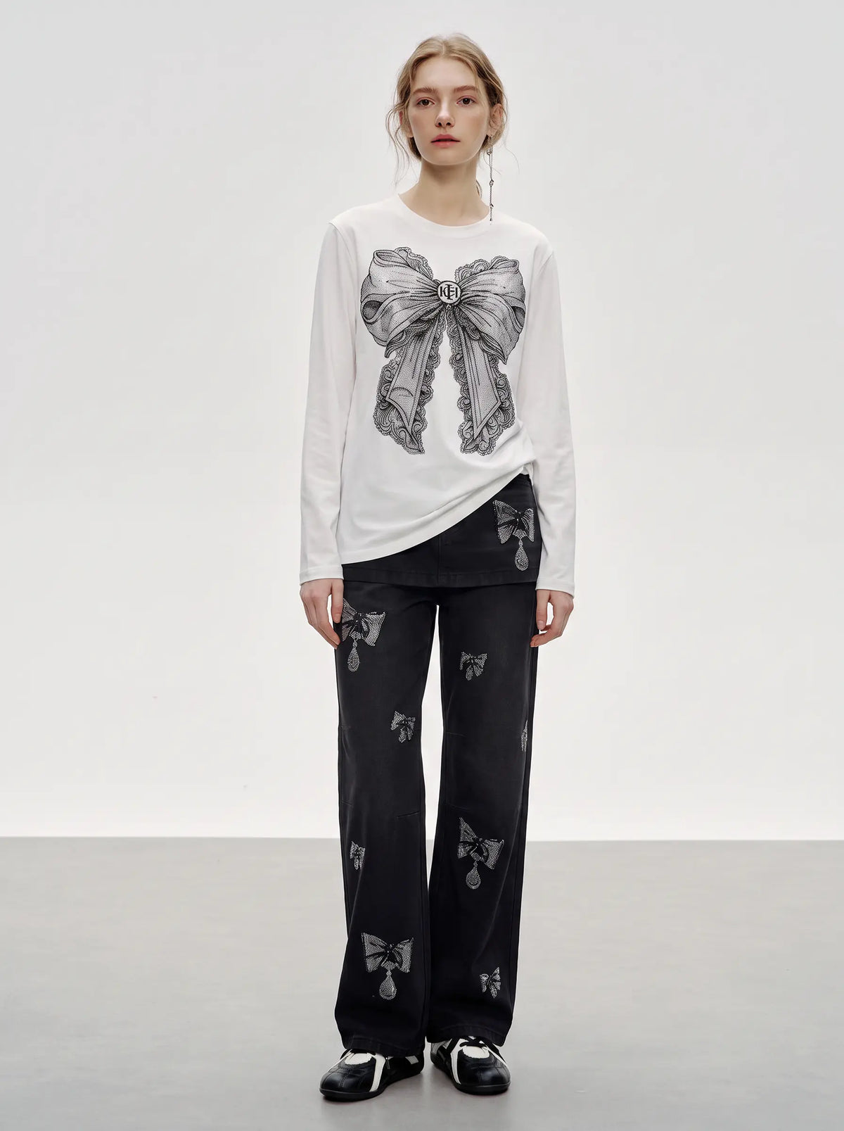 A model stands in a studio wearing a white long-sleeve top with a large gray bow graphic on the front, paired with black wide-leg pants featuring small bow motifs and black-and-white shoes. TCH Rhinestone Bow Graphic Fitted Long Sleeve Top