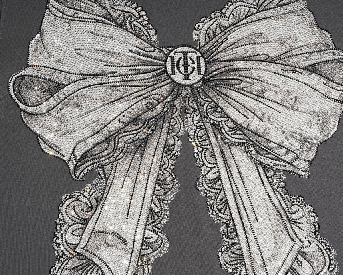 A close-up of a gray garment featuring a large, highly detailed graphic of a white and gray lace bow with a central circular monogram logo. TCH Rhinestone Bow Graphic Fitted Long Sleeve Top