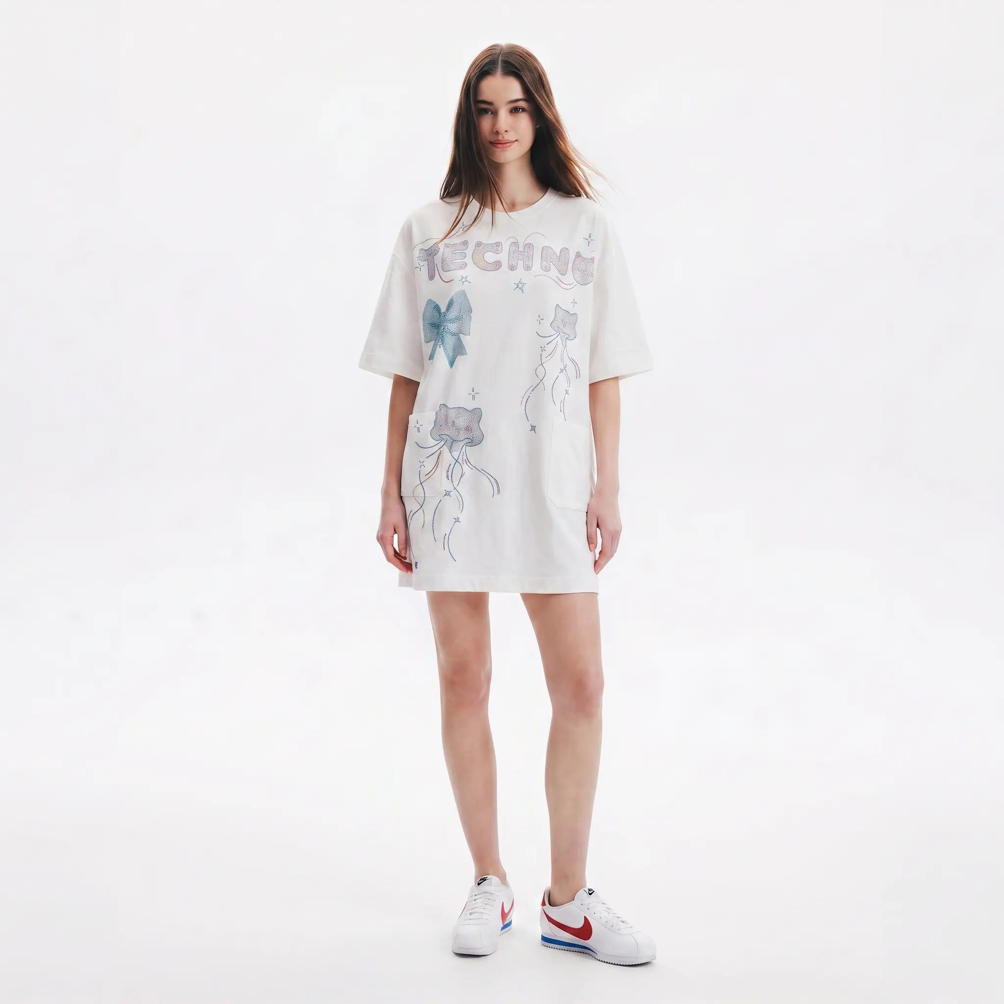 A woman modeling an oversized white graphic T-shirt dress with blue and gray butterfly designs and the word 'TECHNE' across the chest, paired with white sneakers, on a seamless white studio background. TCH Rhinestone Bow Graphic Oversized T-Shirt Dress