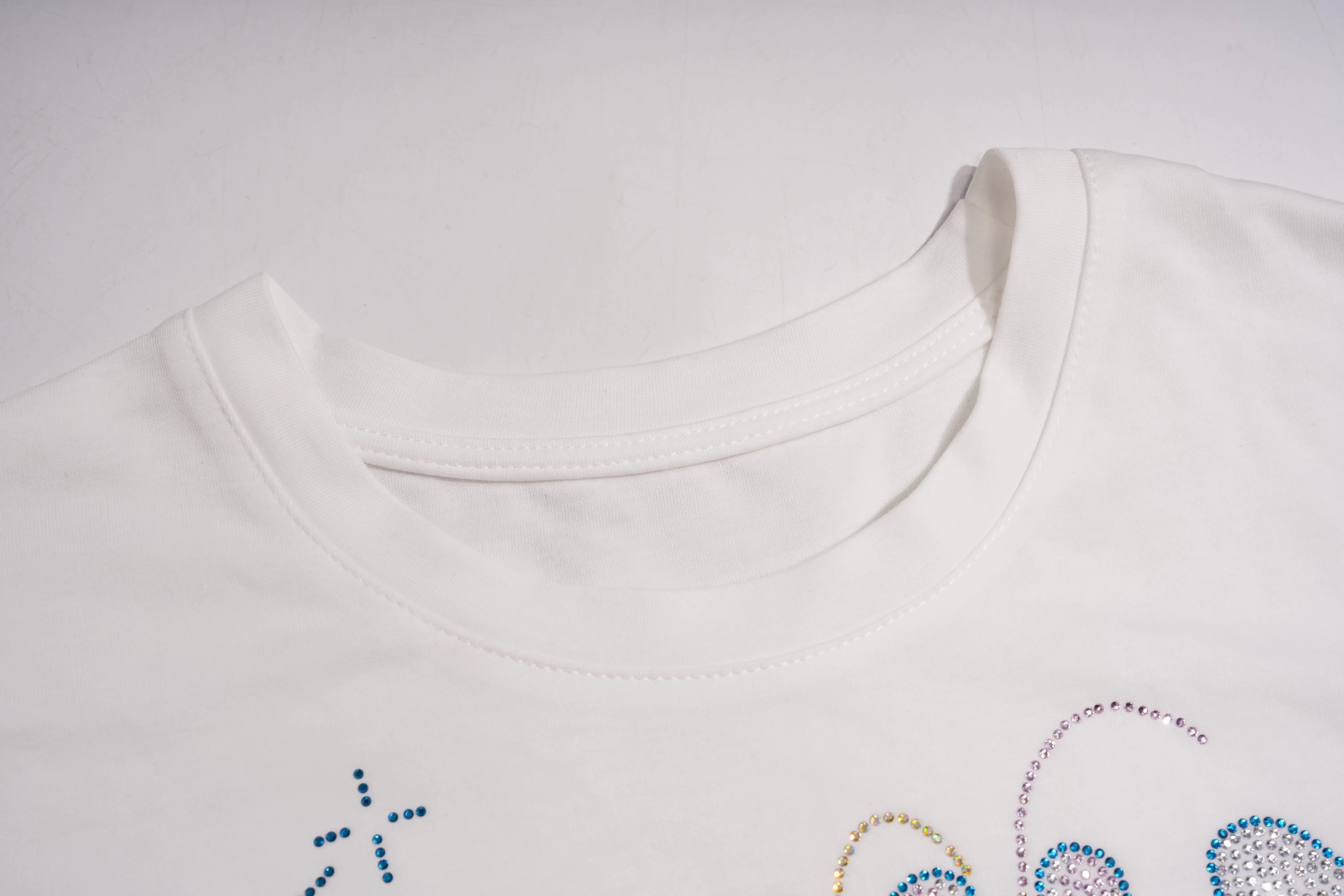 Close-up of a white crewneck t-shirt showing the collar construction and the edge of colorful rhinestone embroidery on the front. TCH Rhinestone Bow Graphic Oversized T-Shirt Dress