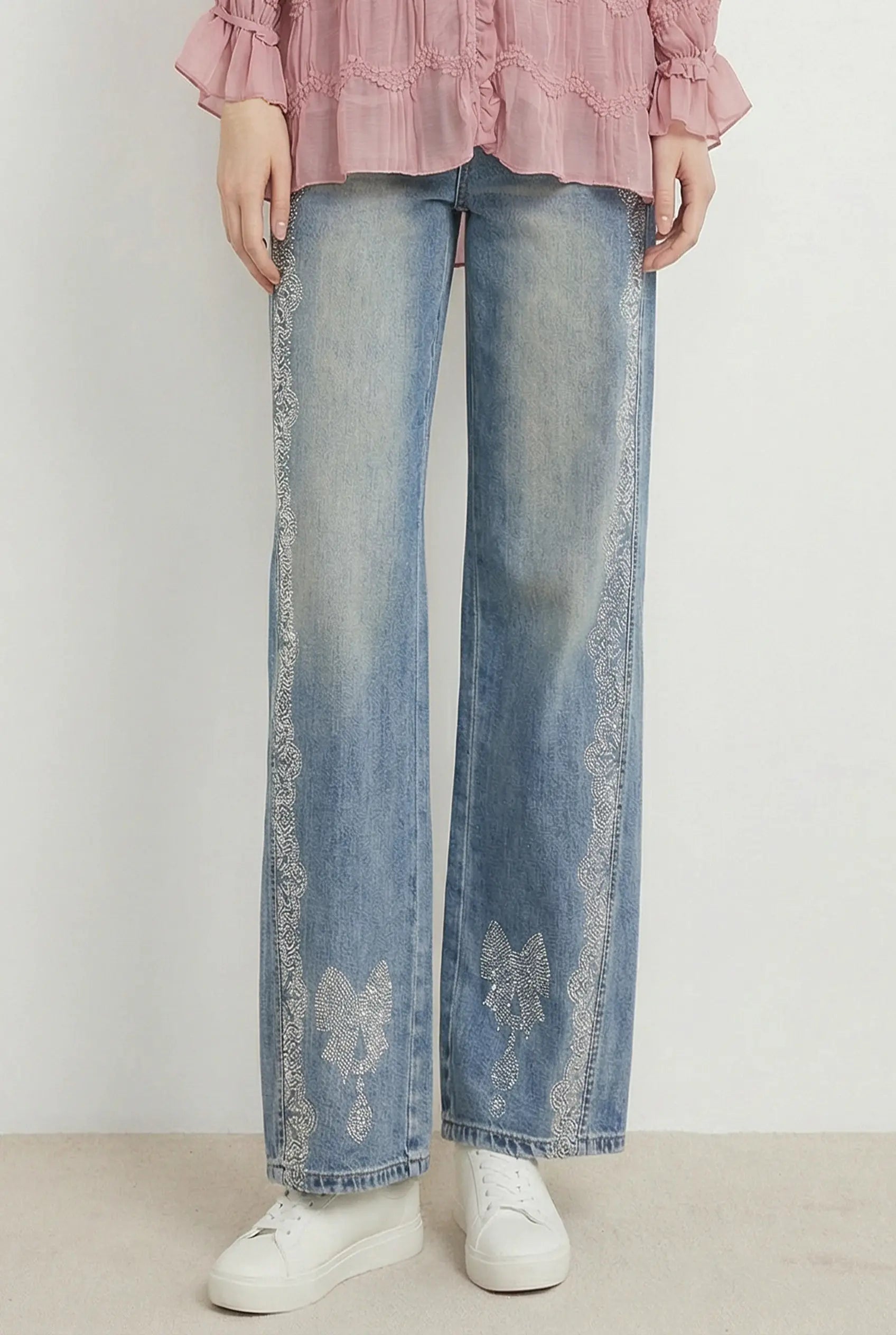 A person wearing light blue wide-leg jeans with white embroidered lace-like detailing down the sides and near the hems, paired with a pink ruffled blouse and white sneakers against a plain light background. TCH Rhinestone Bow Lace Side Panel Wide Leg Jeans