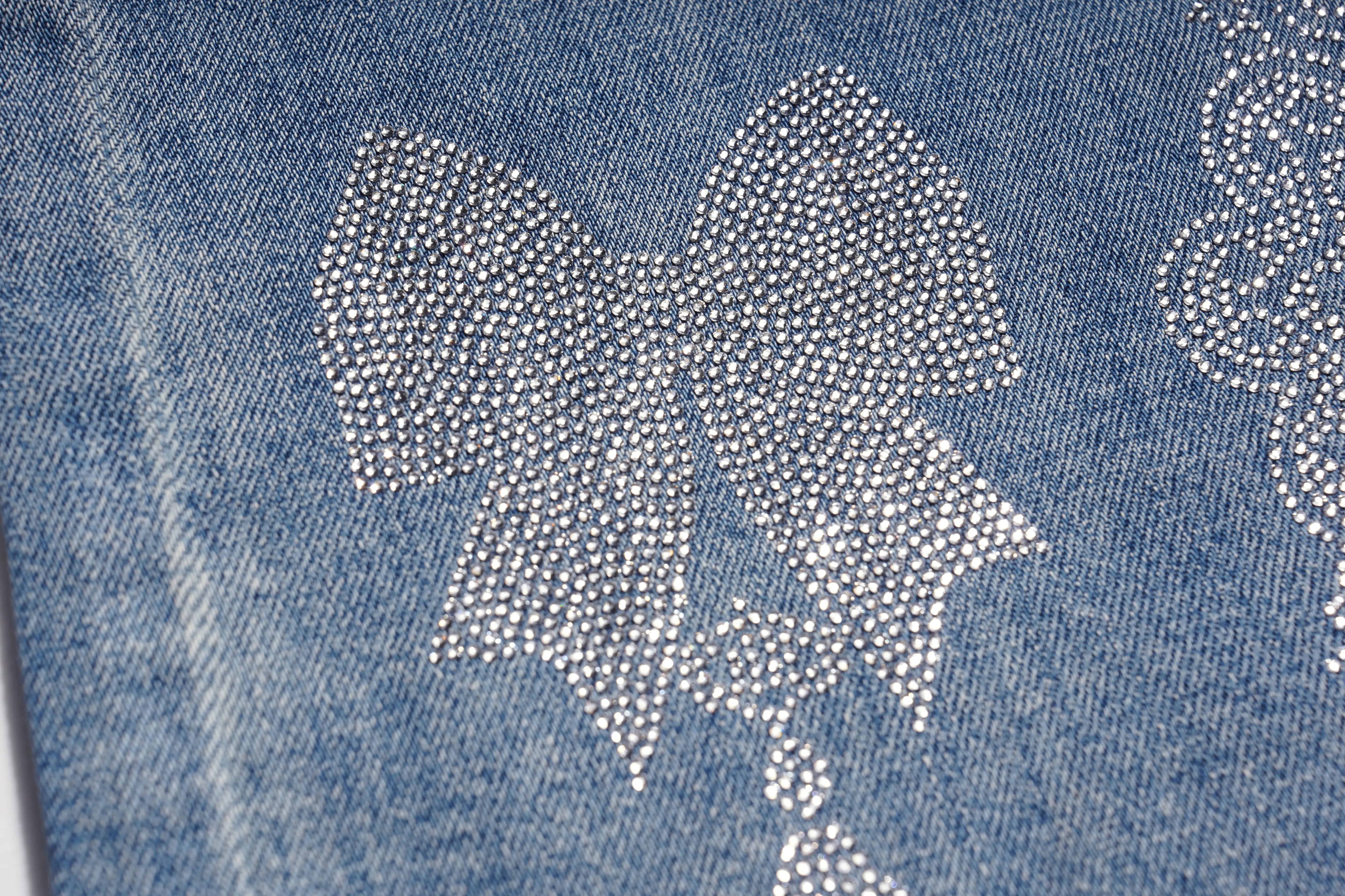 Close-up of a blue denim fabric with a large bow-shaped embellishment made of many small silver rhinestones, and part of another rhinestone motif visible at the right edge. TCH Rhinestone Bow Lace Side Panel Wide Leg Jeans
