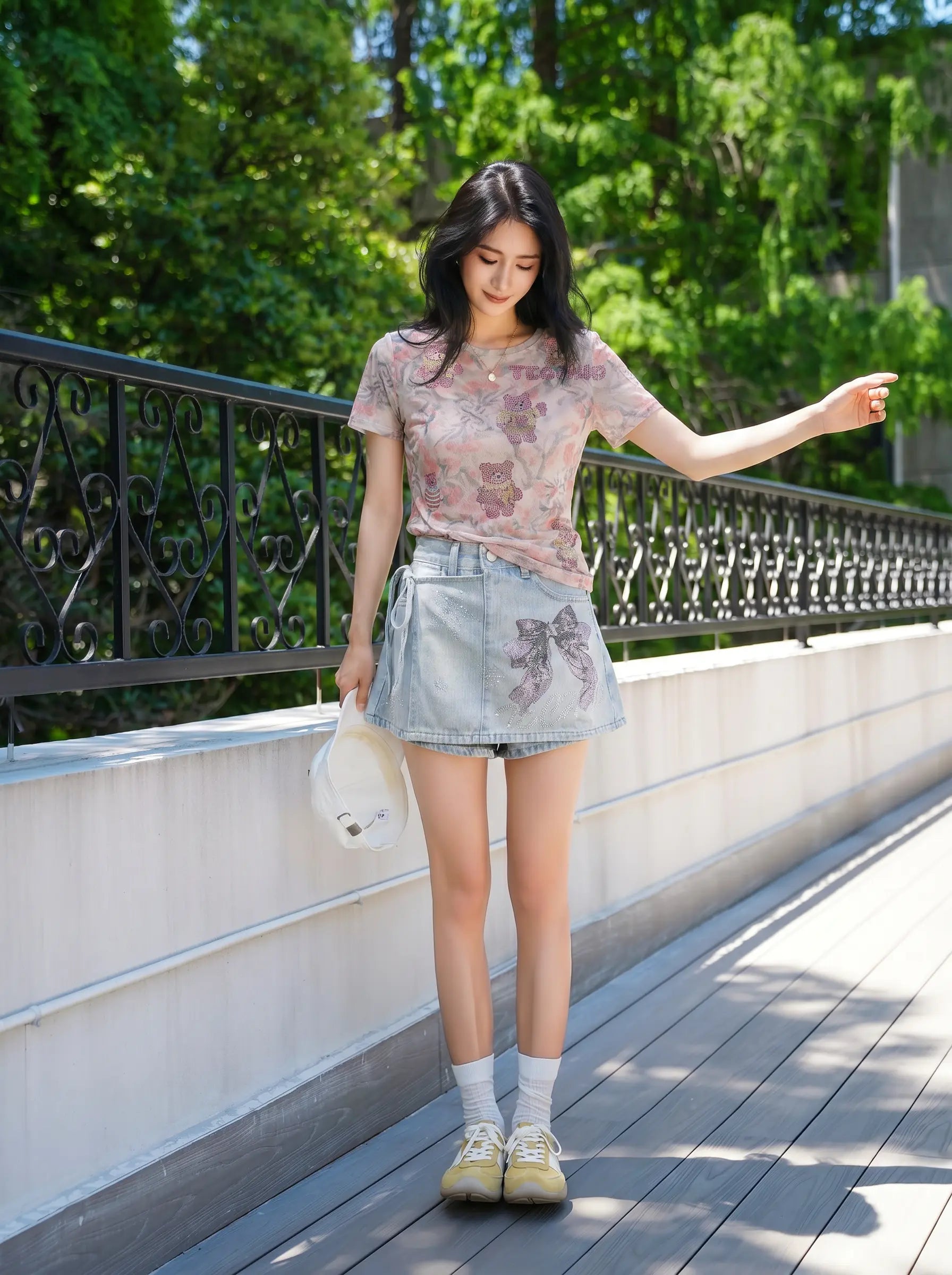 A woman stands on an outdoor wooden walkway beside a railing, wearing a light pink floral short-sleeve T-shirt, a light blue denim mini skirt, white socks, and beige sneakers, while holding a white hat. TCH Rhinestone Bow Light Wash Denim Shorts