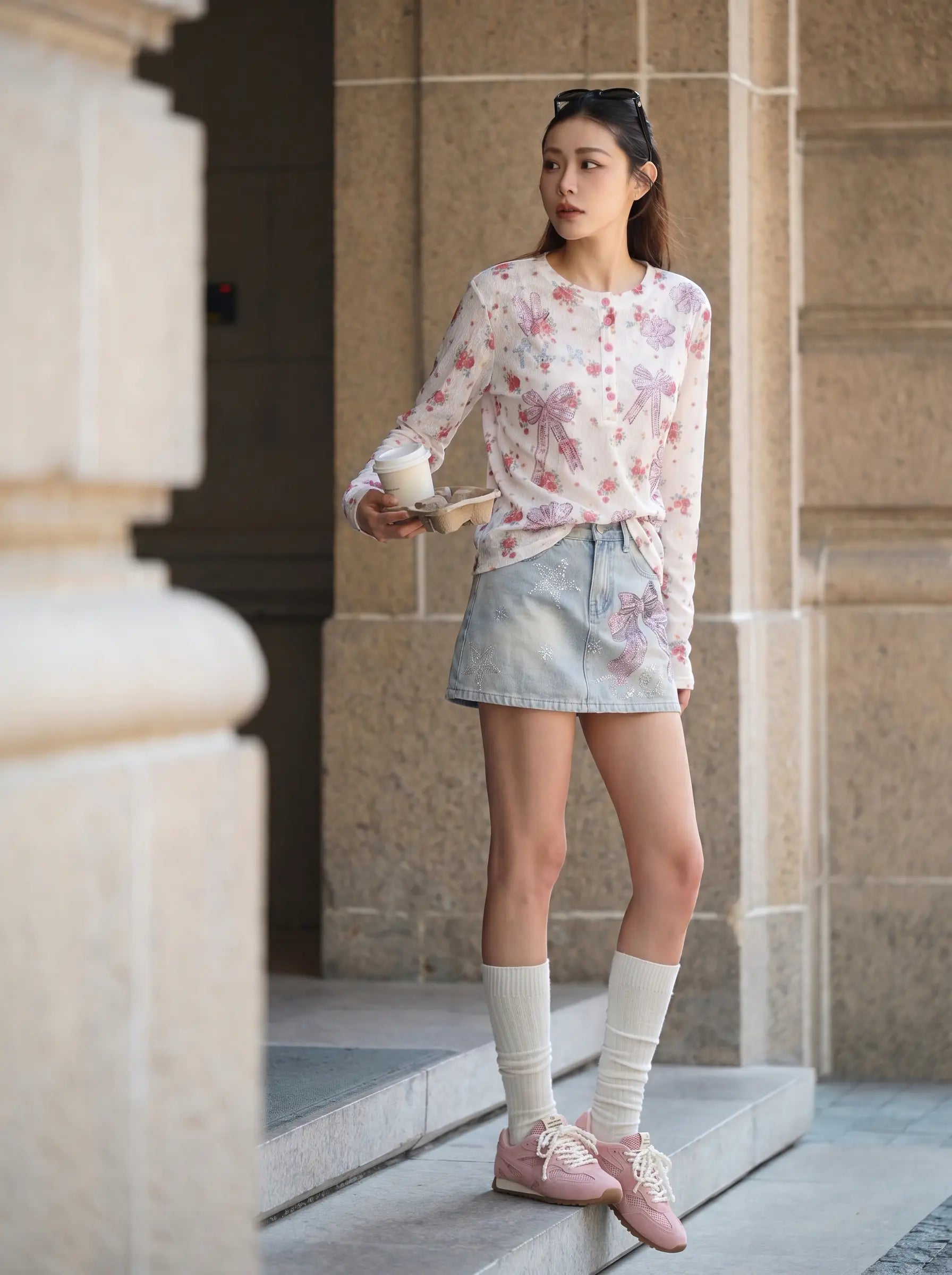 A woman stands on stone steps wearing a light pink floral long-sleeve top, a light blue denim mini skirt, white knee-high socks, and pink sneakers, holding a takeaway coffee cup. TCH Rhinestone Bow Light Wash Denim Shorts