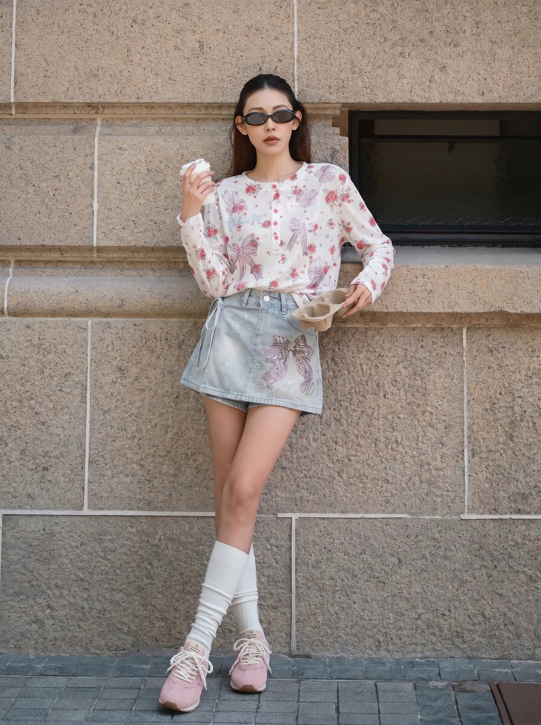 A woman stands against a stone wall wearing a long-sleeve white floral top, a light blue denim mini skirt with a pink floral patch, white knee-high socks, and pink sneakers. TCH Rhinestone Bow Light Wash Denim Shorts