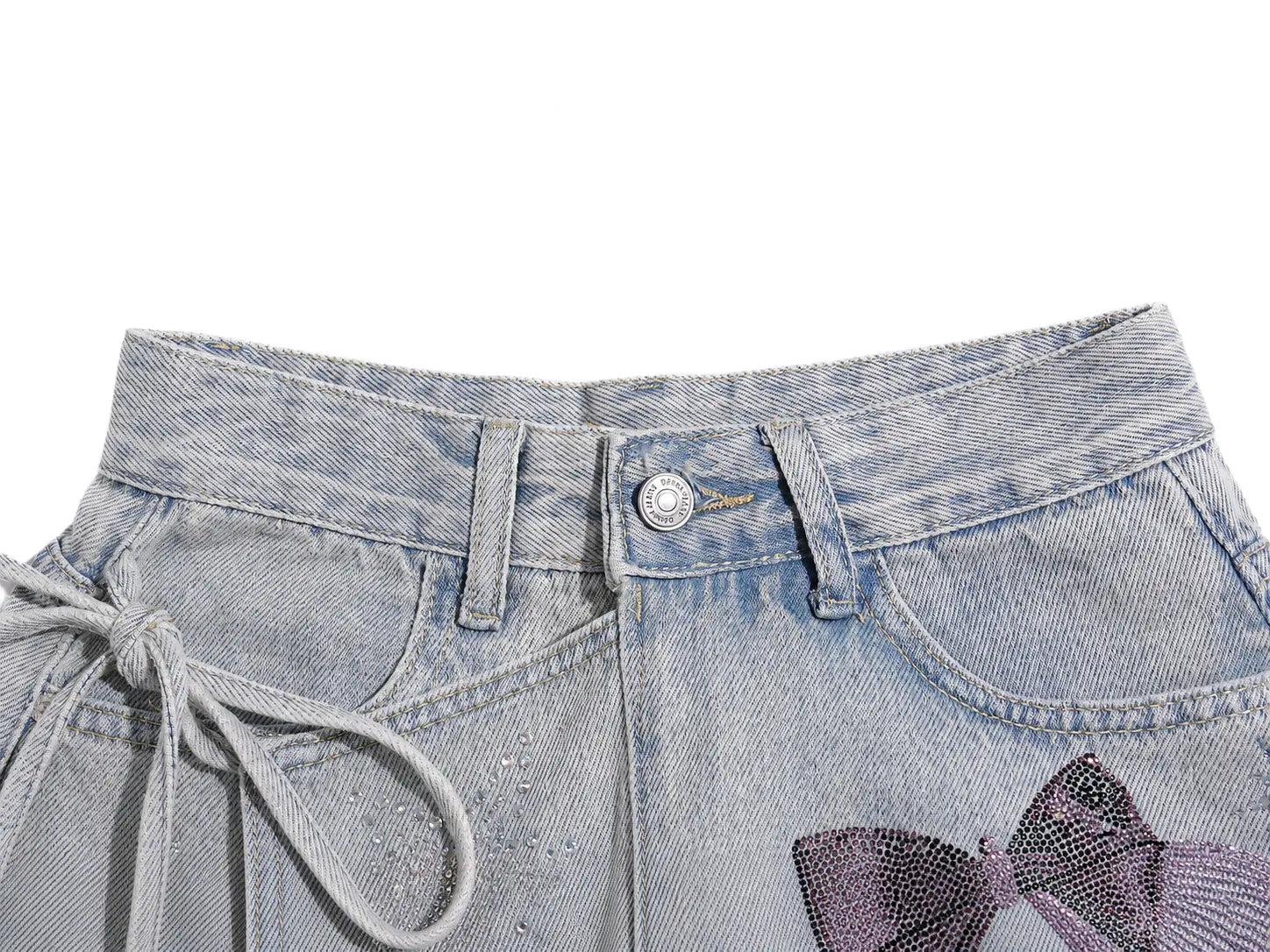 Close-up of a pair of light-wash denim shorts showing the waistband, button closure, side drawstring, and a purple rhinestone bow embellishment on the front. TCH Rhinestone Bow Light Wash Denim Shorts