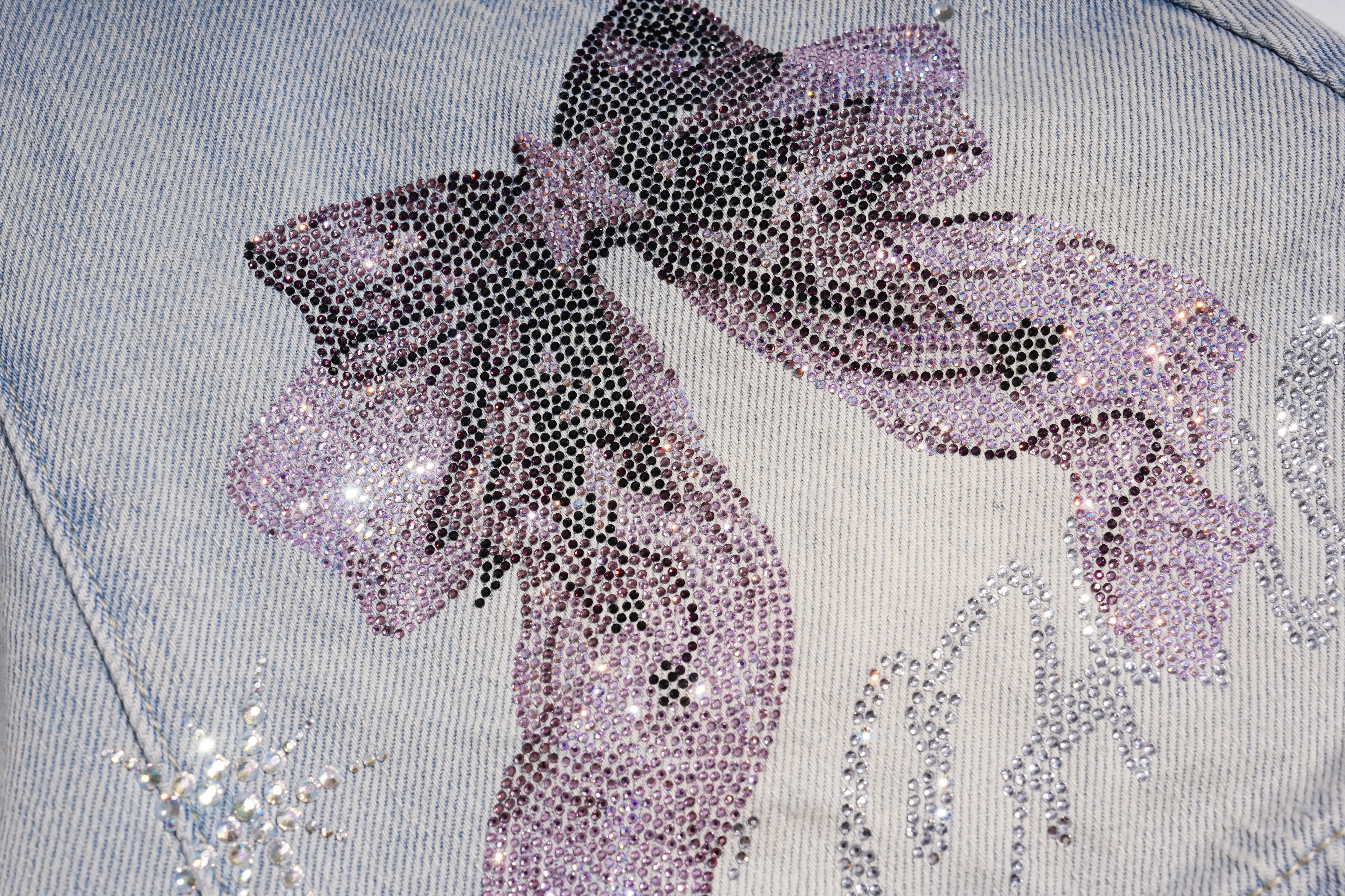 Close-up of a light blue denim garment featuring a large bow embellishment made of black and lavender-pink rhinestones, with silver rhinestone details nearby. TCH Rhinestone Bow Light Wash Denim Shorts