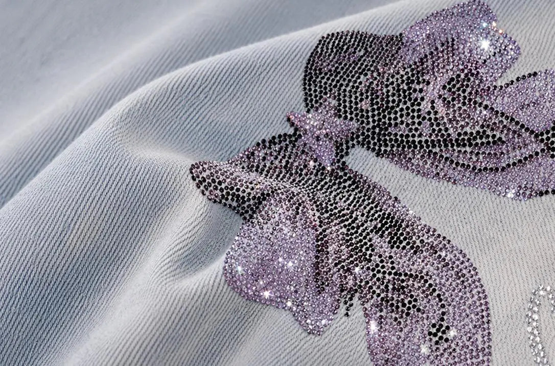 A close-up of a light gray textured fabric featuring a large, ornate bow embellished with dense rows of sparkling lavender and silver rhinestones. TCH Rhinestone Bow Light Wash Denim Shorts