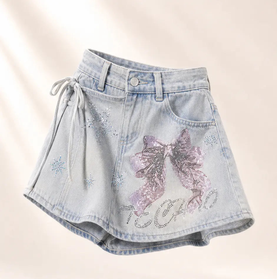 Light-wash denim shorts with a large sequined purple bow on the front, rhinestone star details, a side drawstring, and 'TOO COLD' written in rhinestones near the hem, shown on a warm off-white background. TCH Rhinestone Bow Light Wash Denim Shorts