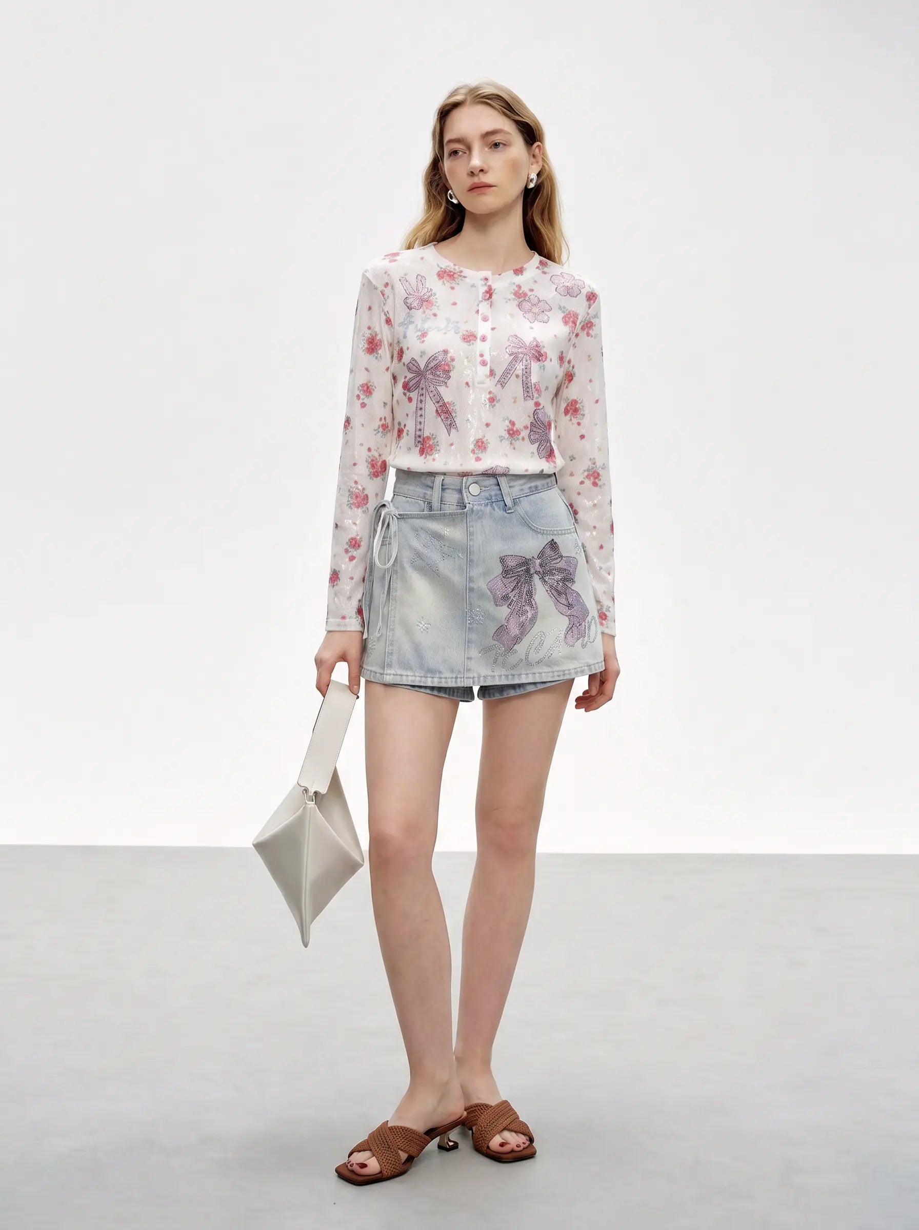 A model stands in a studio wearing a long-sleeve white button-up blouse with a pink and purple floral print, a light-wash denim mini skirt with a purple bow print, and brown slide sandals, while holding a small white handbag. TCH Rhinestone Bow Light Wash Denim Shorts