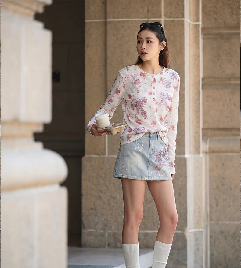 A woman stands outdoors in front of a stone building, wearing a long-sleeve white blouse with a pink and purple floral print and a light denim mini skirt. She holds a takeaway coffee cup and a small card, with white socks pulled up to mid-calf. TCH Rhinestone Bow Light Wash Denim Shorts