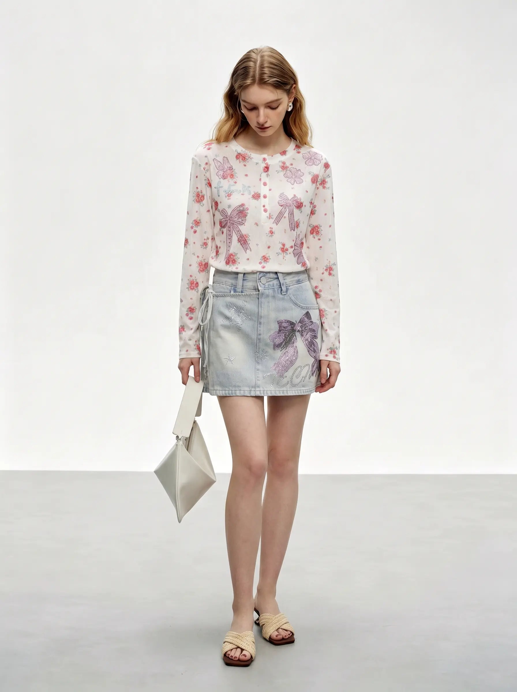 A model stands in a studio wearing a sheer long-sleeve button-up blouse with a pink and purple floral print over a white base, paired with a light-wash denim mini skirt featuring a purple floral print, holding a small white handbag. TCH Rhinestone Bow Light Wash Denim Shorts