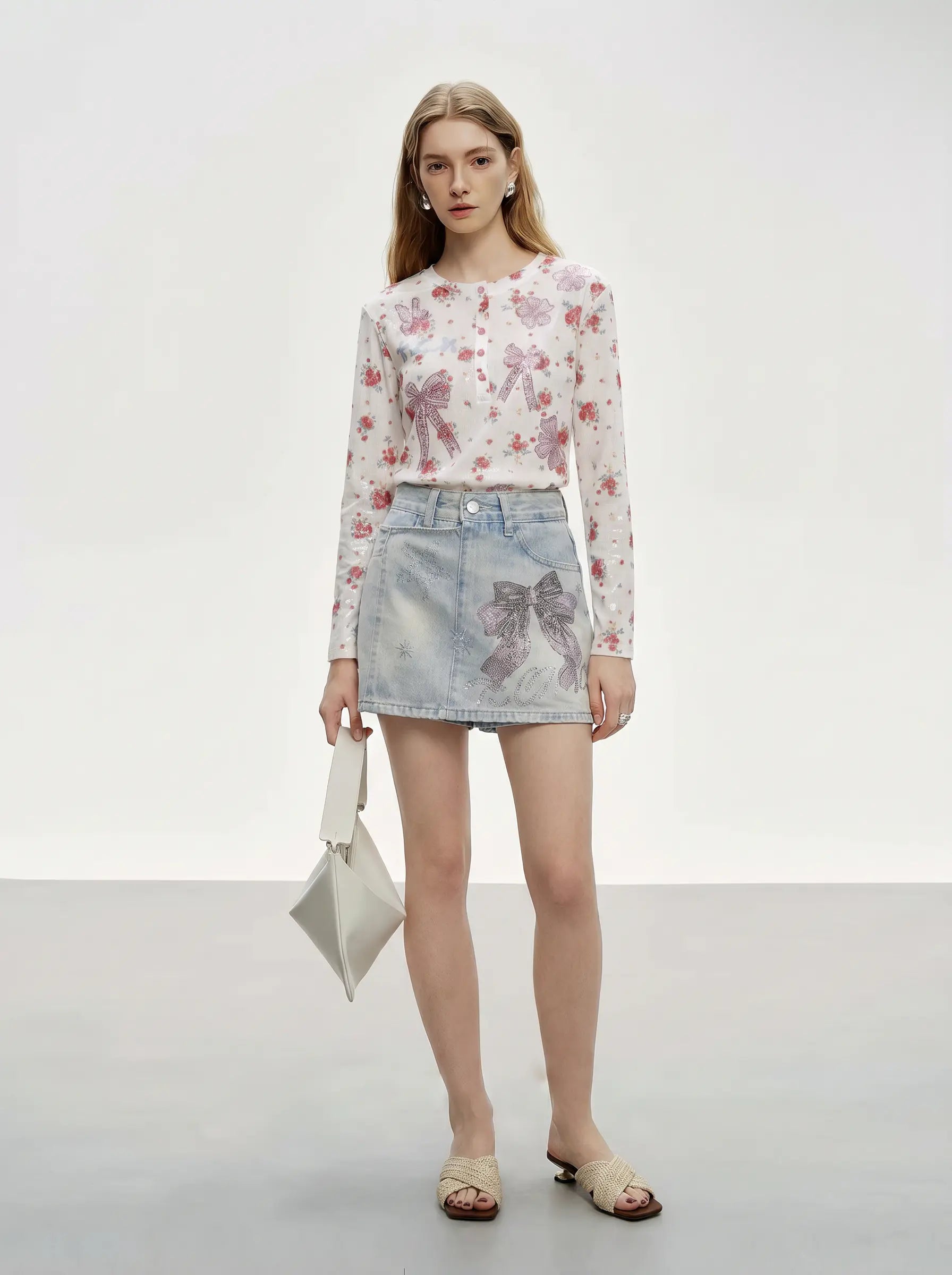 A model stands in a studio wearing a sheer long-sleeve top with a pink and red floral print over a white base, paired with a light-wash denim mini skirt featuring a bow print, holding a small white handbag. TCH Rhinestone Bow Light Wash Denim Shorts