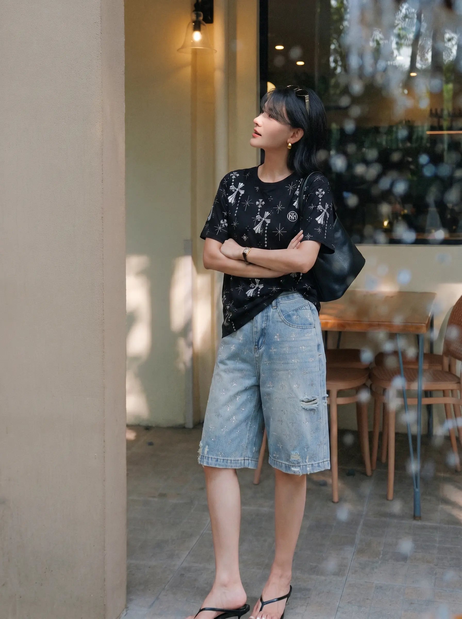 A woman stands outdoors near a cafe doorway wearing a black short-sleeve t-shirt with white floral embroidery, light-wash denim shorts, and black sandals. TCH Rhinestone Bow Star Pattern Black T-Shirt