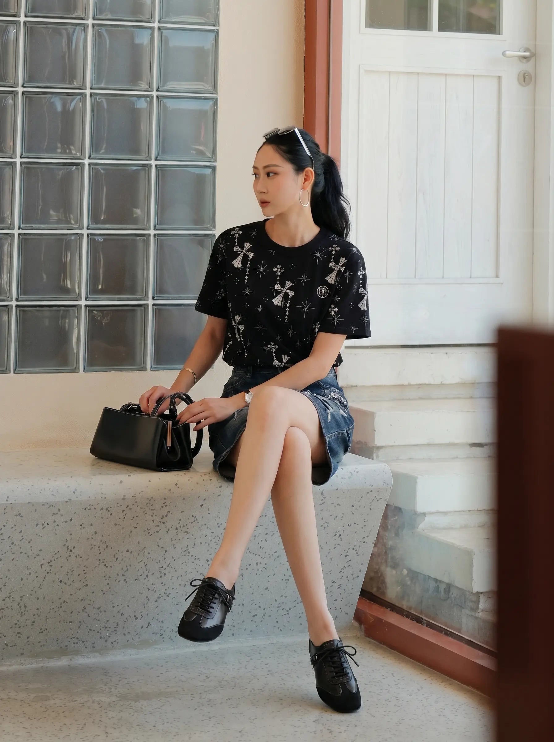 A woman sits on a concrete bench in front of a glass block wall and white door, wearing a black short-sleeve t-shirt with a white cross pattern, denim shorts, and black lace-up shoes, while holding a black handbag. TCH Rhinestone Bow Star Pattern Black T-Shirt