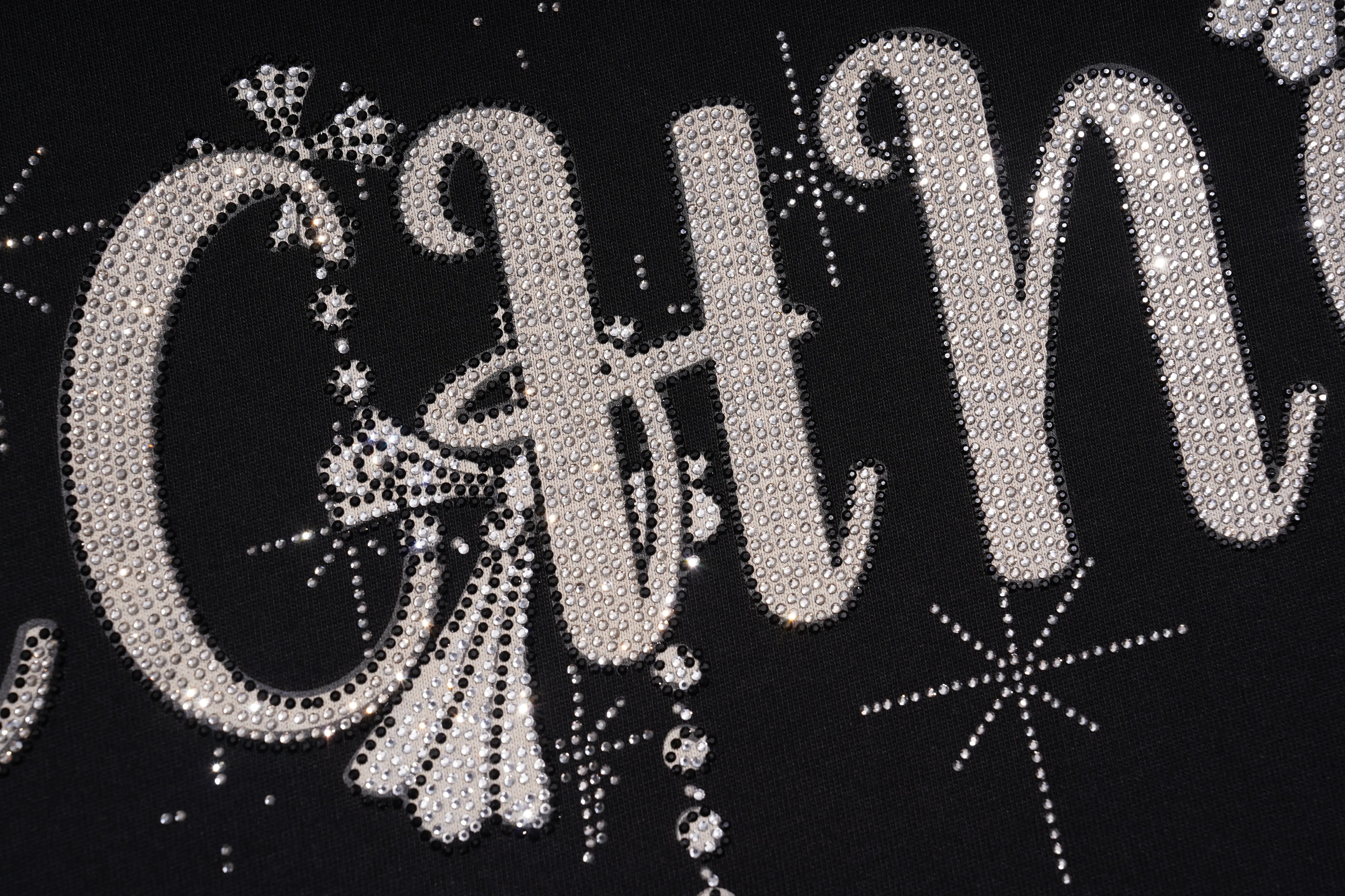 A close-up of a black garment featuring large silver rhinestone lettering and decorative snowflake embellishments. TCH Rhinestone Bow Star Pattern Black T-Shirt