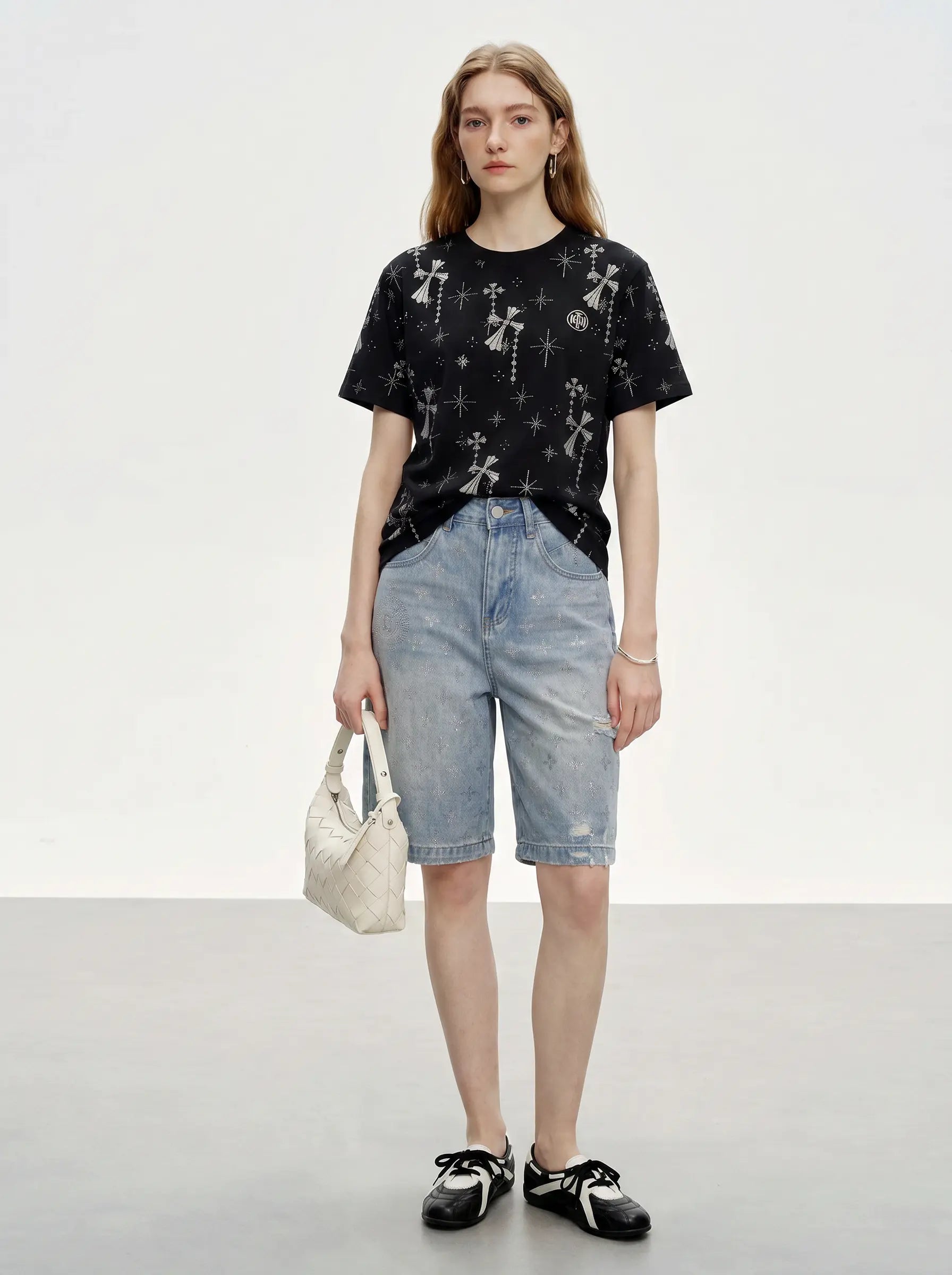 A model stands against a light gray studio background wearing a black short-sleeve t-shirt with a white graphic print, light-wash denim shorts, black and white loafers, and holding a small white handbag. TCH Rhinestone Bow Star Pattern Black T-Shirt