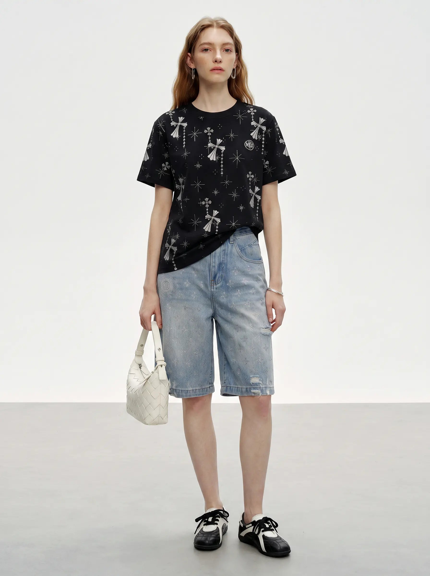 A model stands against a light gray studio background wearing a black short-sleeve T-shirt with a white gothic cross and star pattern, light-wash denim shorts, black-and-white sneakers, and holding a small white handbag. TCH Rhinestone Bow Star Pattern Black T-Shirt