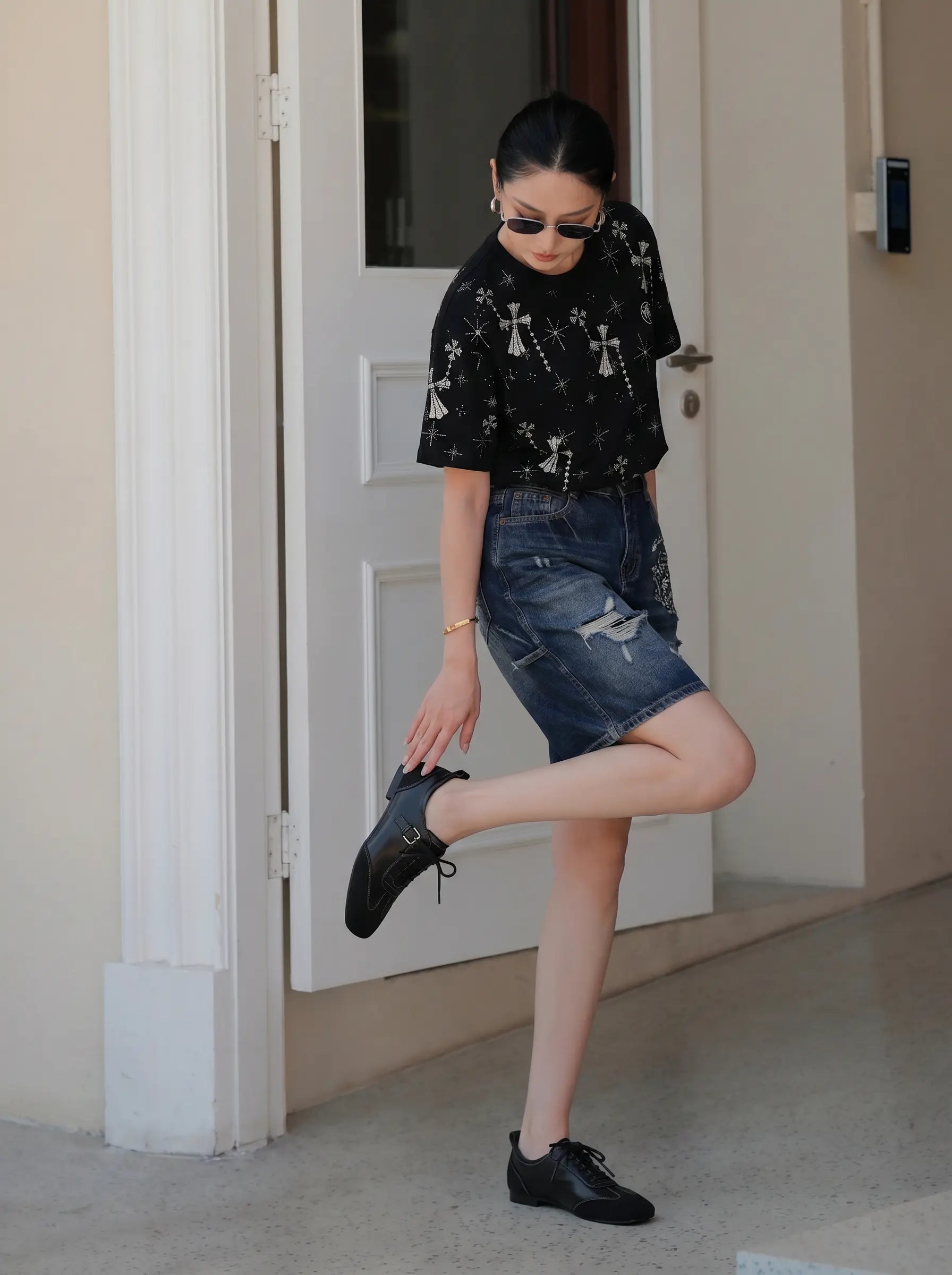A woman posing in front of a white door, wearing a black short-sleeve t-shirt with a white graphic print, a distressed denim mini skirt, and black leather shoes. TCH Rhinestone Bow Star Pattern Black T-Shirt