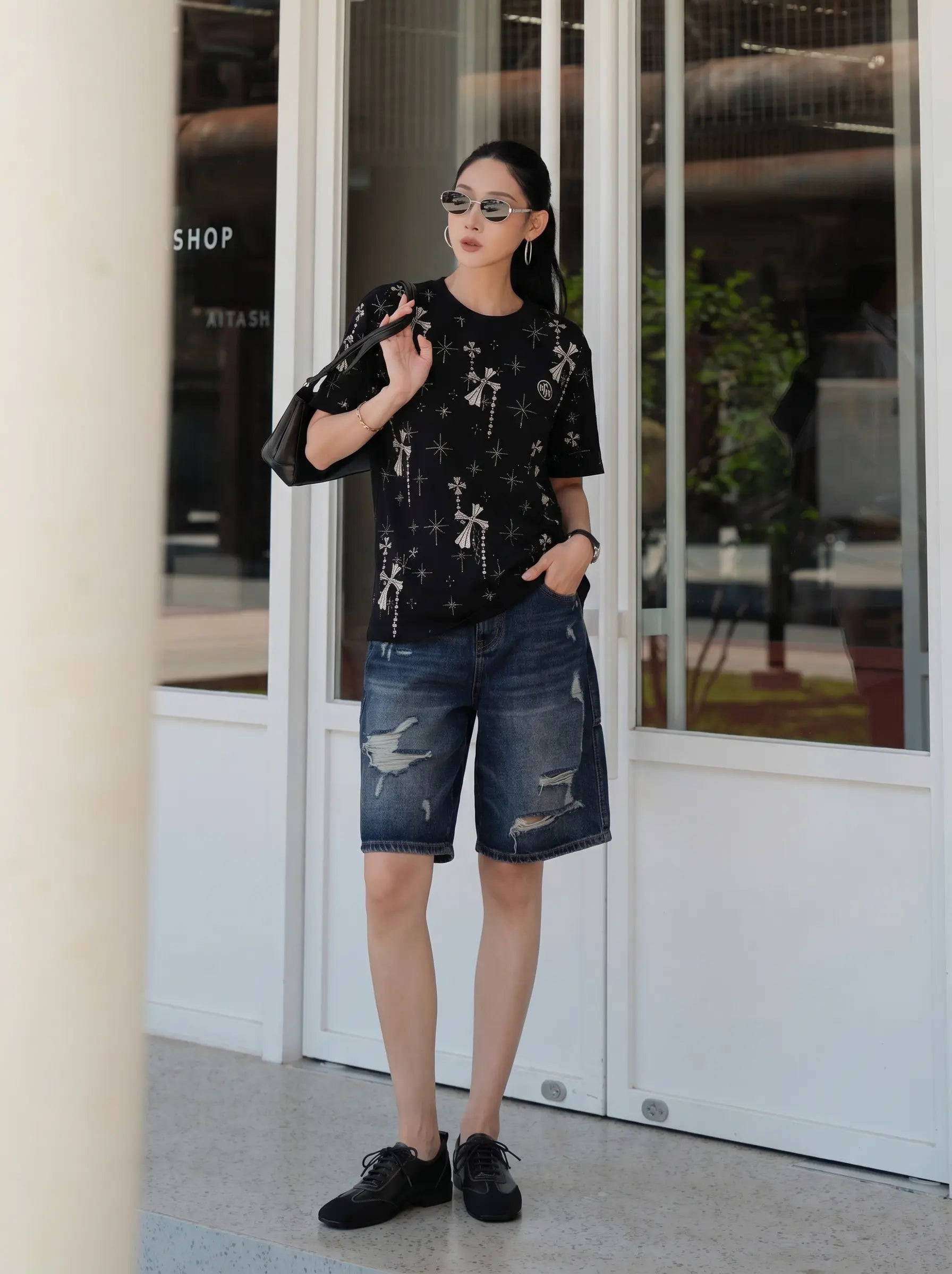 A woman stands in front of a white storefront with glass doors, wearing a black short-sleeve t-shirt with a white cross pattern, distressed blue denim shorts, and black shoes. TCH Rhinestone Bow Star Pattern Black T-Shirt
