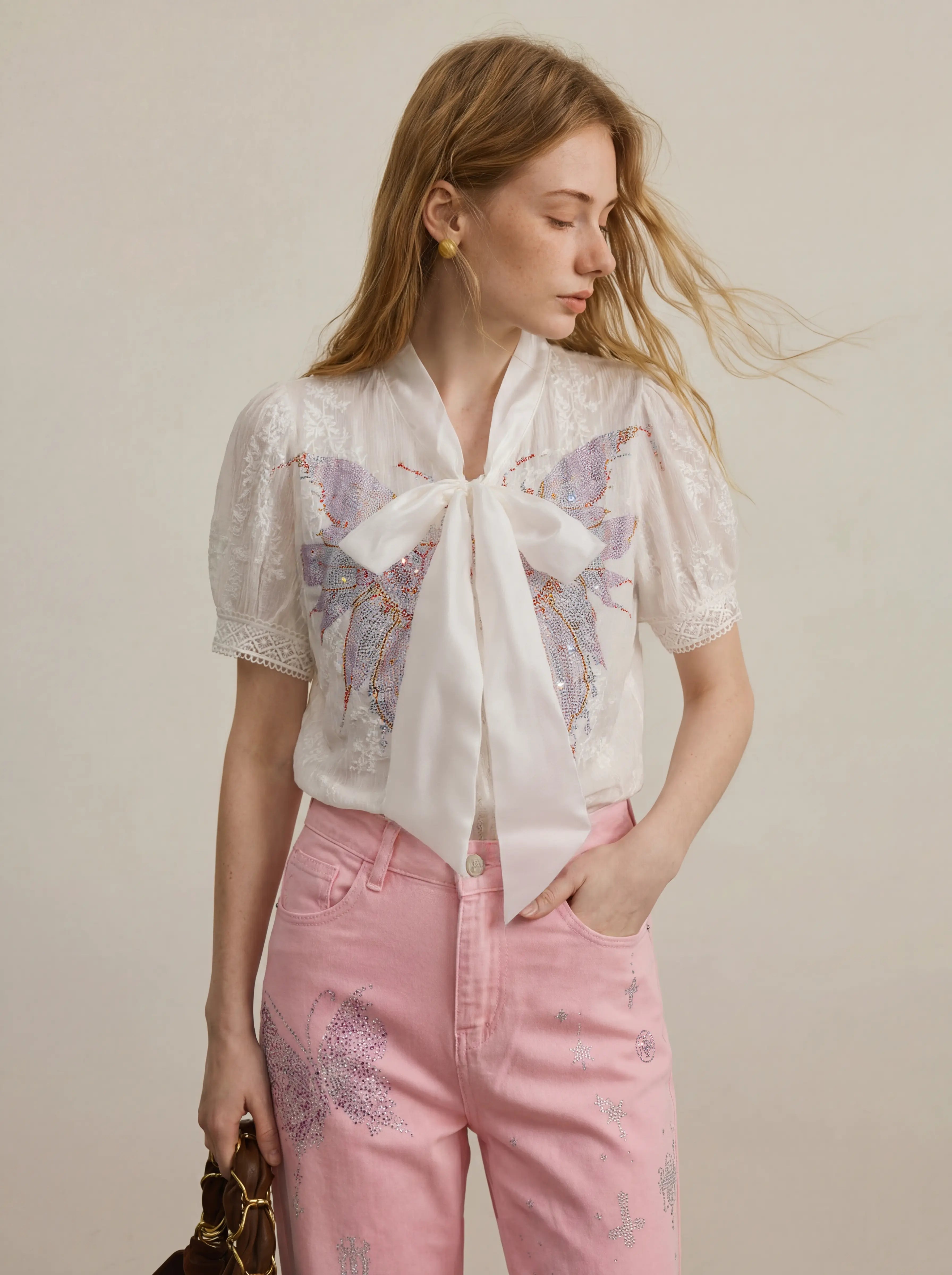 A woman wearing a white short-sleeve lace blouse with a large bow and butterfly embroidery, paired with pink pants, photographed against a plain light beige studio background. TCH Rhinestone Butterfly Bow Lace Blouse