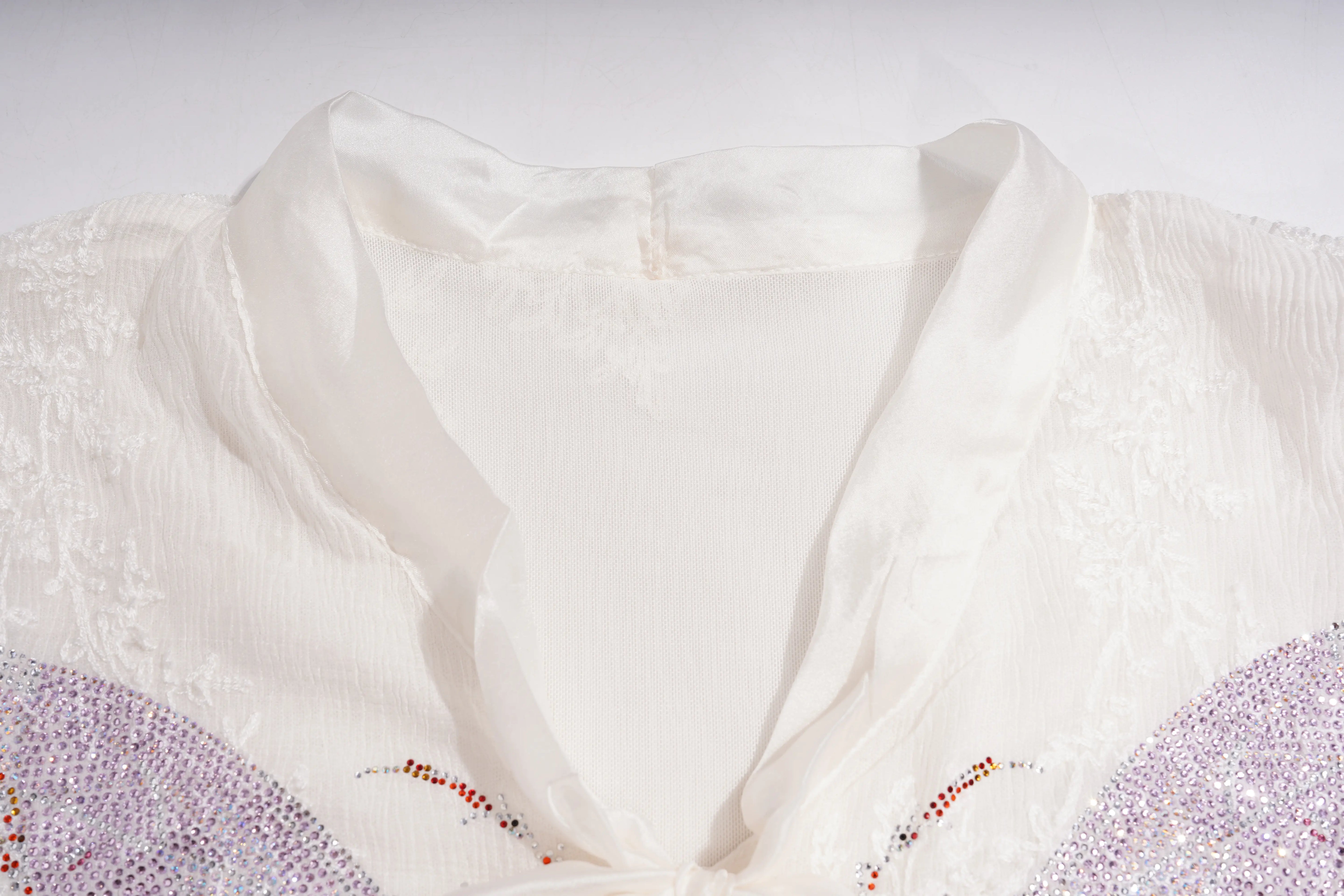 Close-up of a white or ivory sheer mesh garment with embroidered lace detailing and beaded embellishments along the neckline. TCH Rhinestone Butterfly Bow Lace Blouse