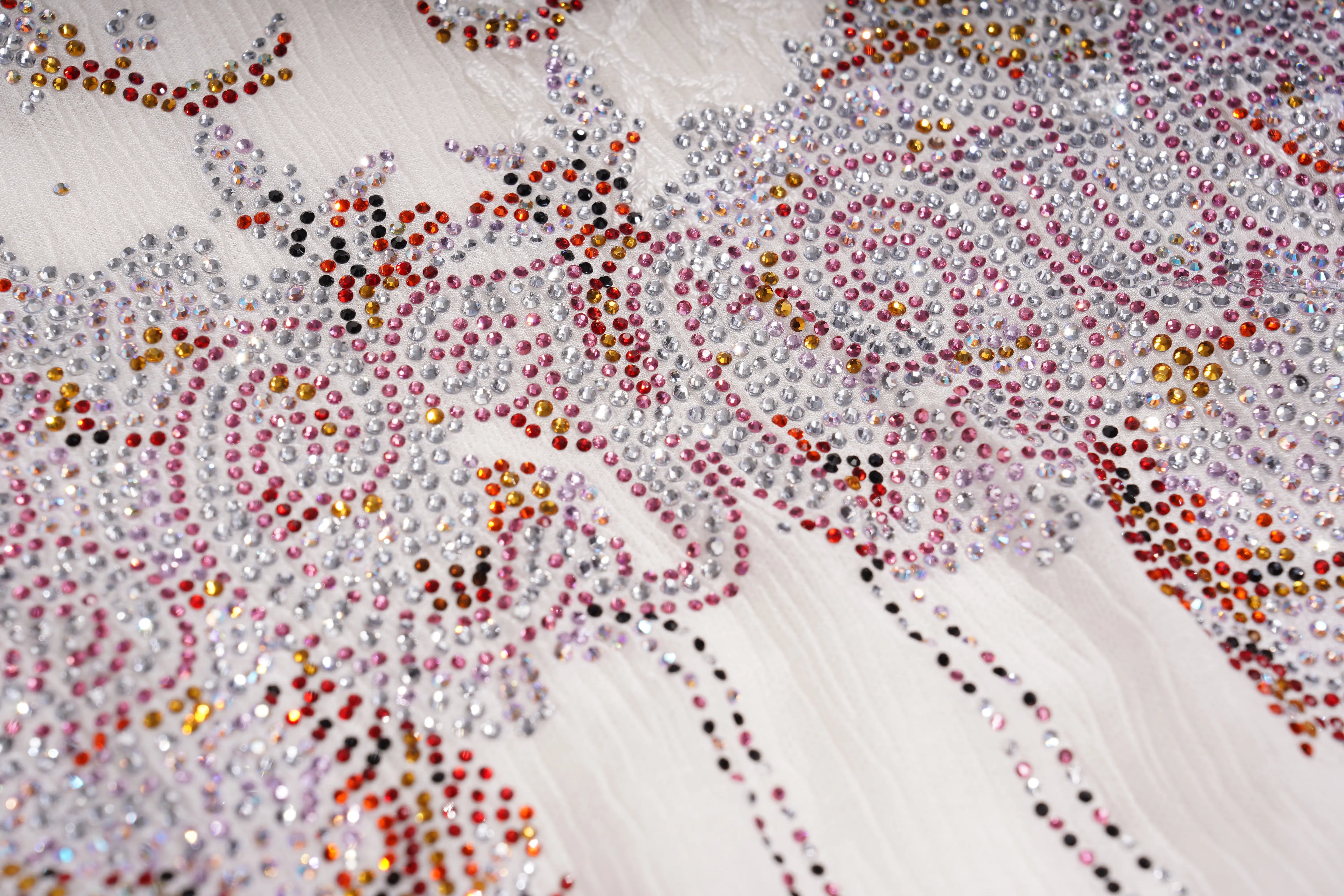 Close-up of a white embellished fabric with swirling floral patterns made of clear rhinestones and rows of pink, red, orange, and black rhinestones. TCH Rhinestone Butterfly Bow Lace Blouse