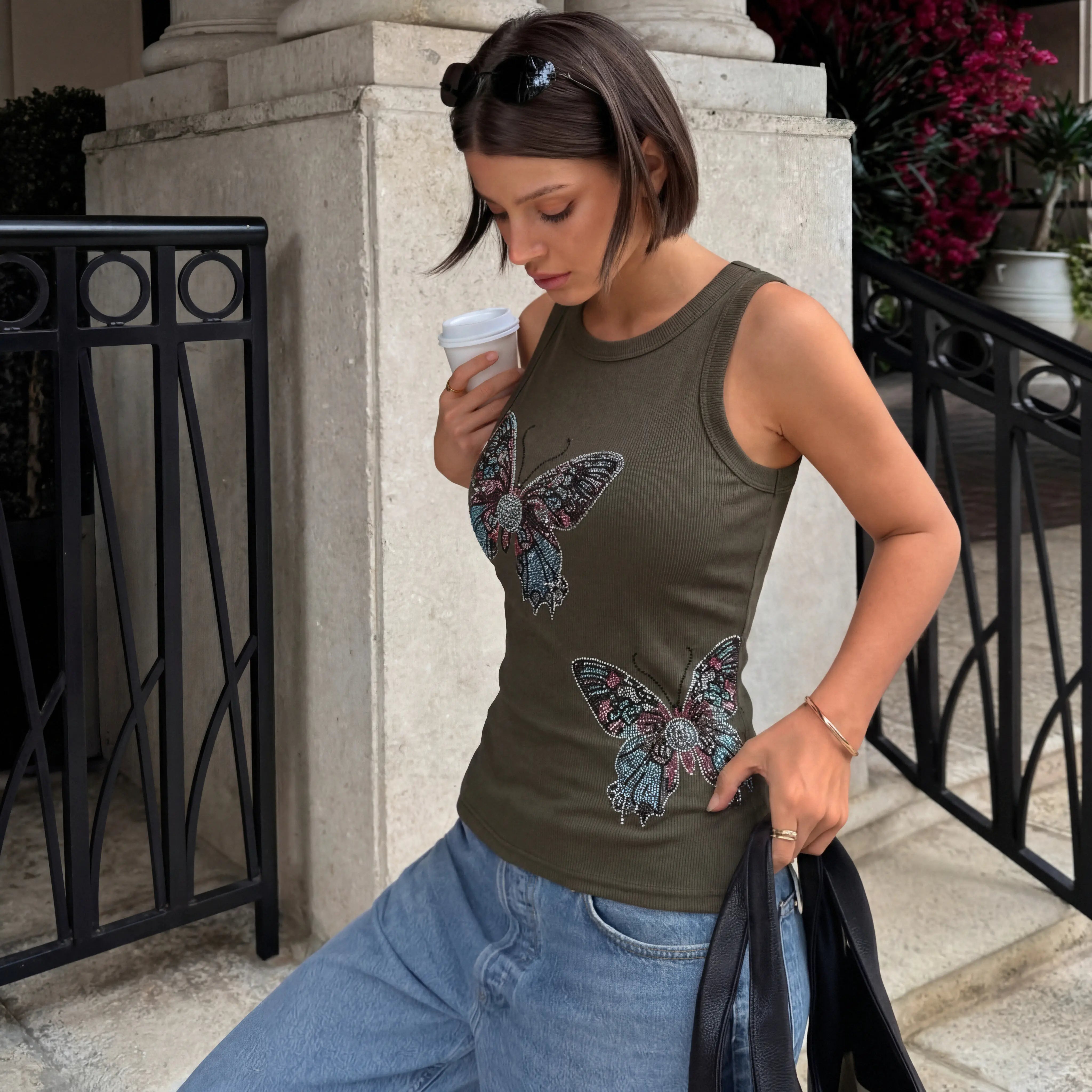 Woman wearing a tank top with butterfly designs, holding a coffee cup outdoors. TCH Rhinestone Butterfly Bow Tank Top