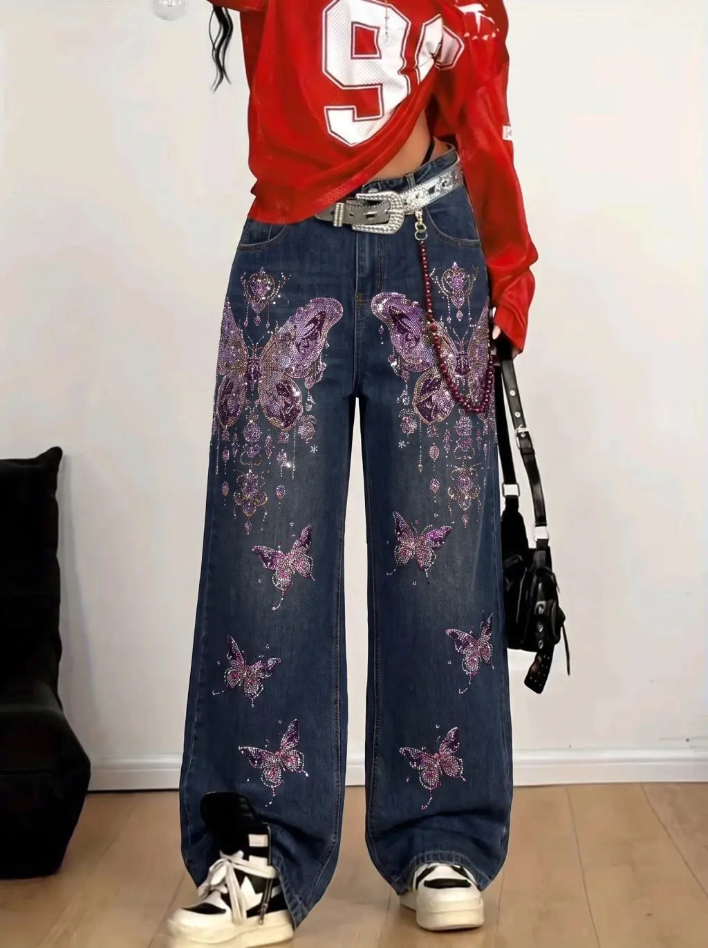 A person wearing wide-leg blue denim jeans with purple butterfly and floral embellishments, paired with a red cropped shirt, silver belt, and white sneakers, standing indoors against a white wall. TCH Rhinestone Butterfly Charm Wide-Leg Denim Jeans