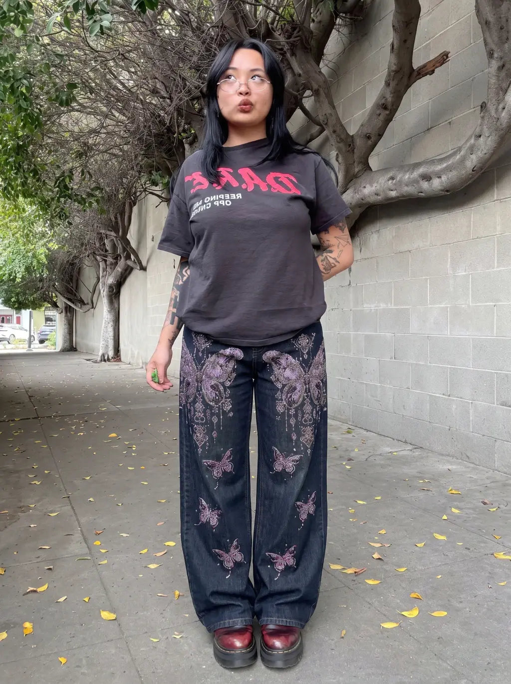 A person standing outdoors on a sidewalk wearing a dark charcoal graphic t-shirt with pink lettering, dark blue wide-leg jeans with pink butterfly and floral prints, and dark red shoes. TCH Rhinestone Butterfly Charm Wide-Leg Denim Jeans